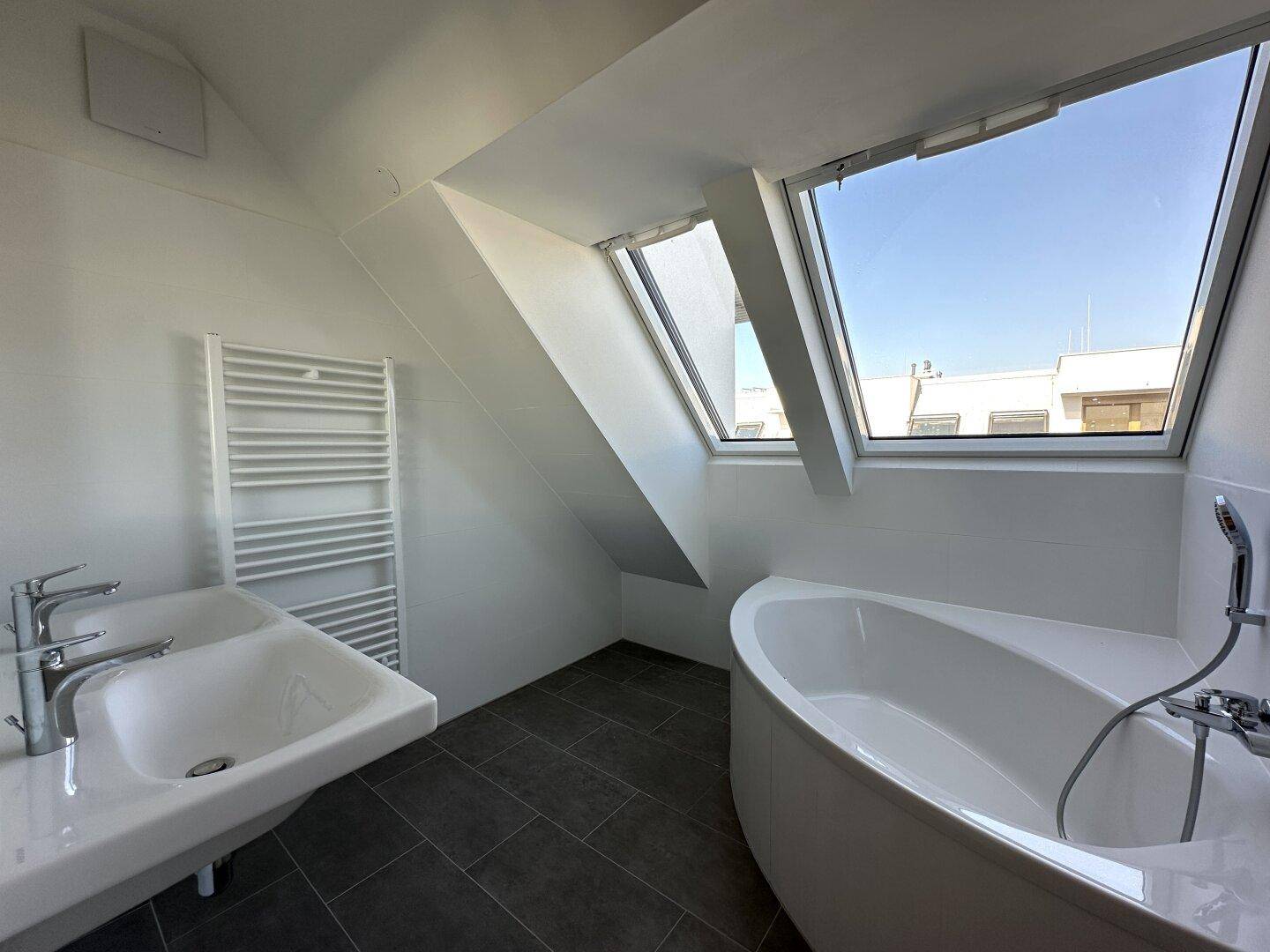 Modernes Badezimmer mit Eckbadewanne, Doppelwaschbecken und großen Dachfenstern unter einer Schrägdecke.
