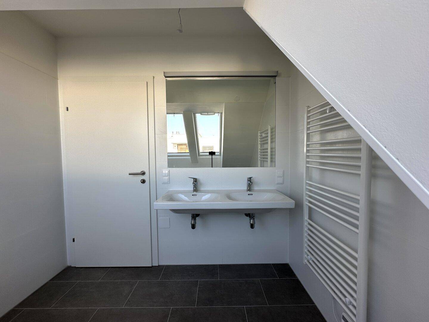 Helles Badezimmer mit Doppelwaschbecken, großem Spiegel und beheiztem Handtuchhalter unter einer Schrägdecke.
