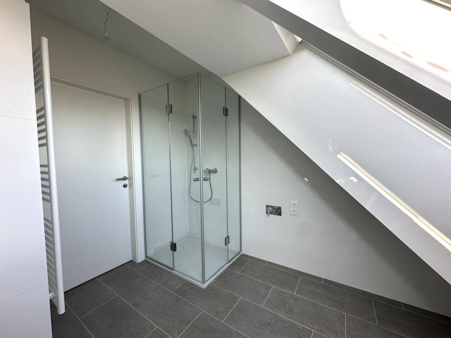 Modernes Badezimmer mit Glasdusche und dunklen Bodenfliesen unter einer Dachschräge.