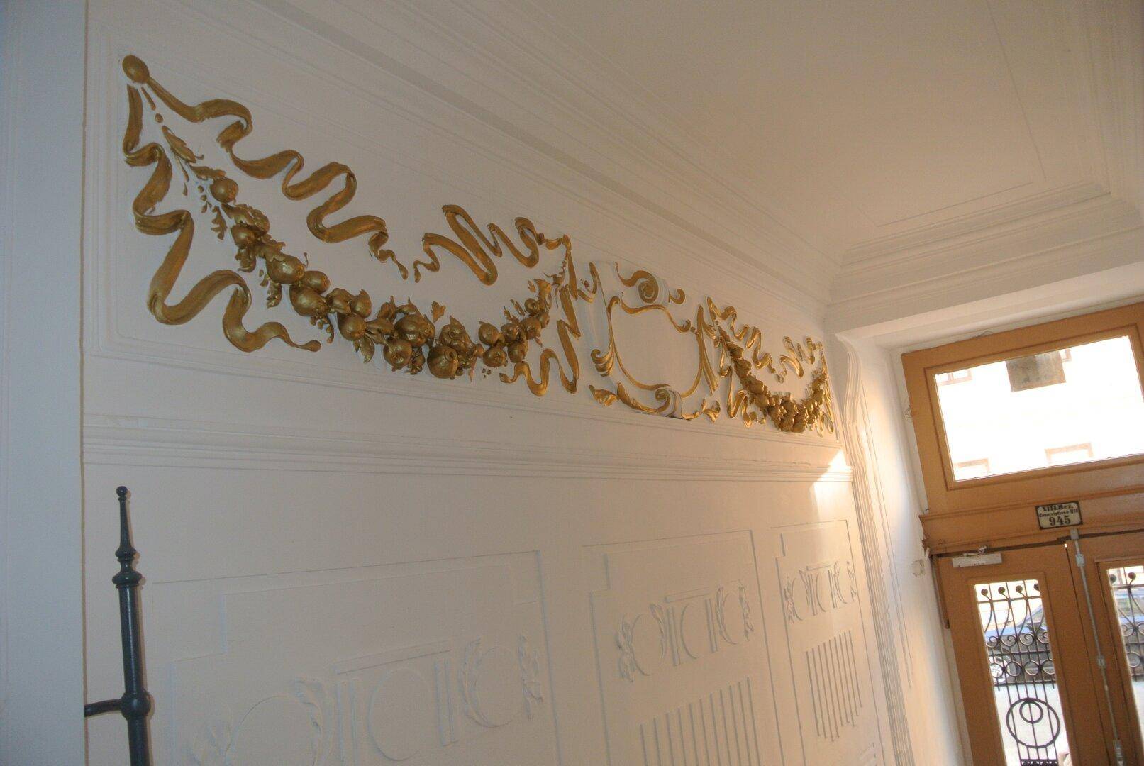 Detailaufnahme von aufwendigem goldenem Stuckdekor an einer weißen Wand im Flurbereich, der Eleganz verleiht.