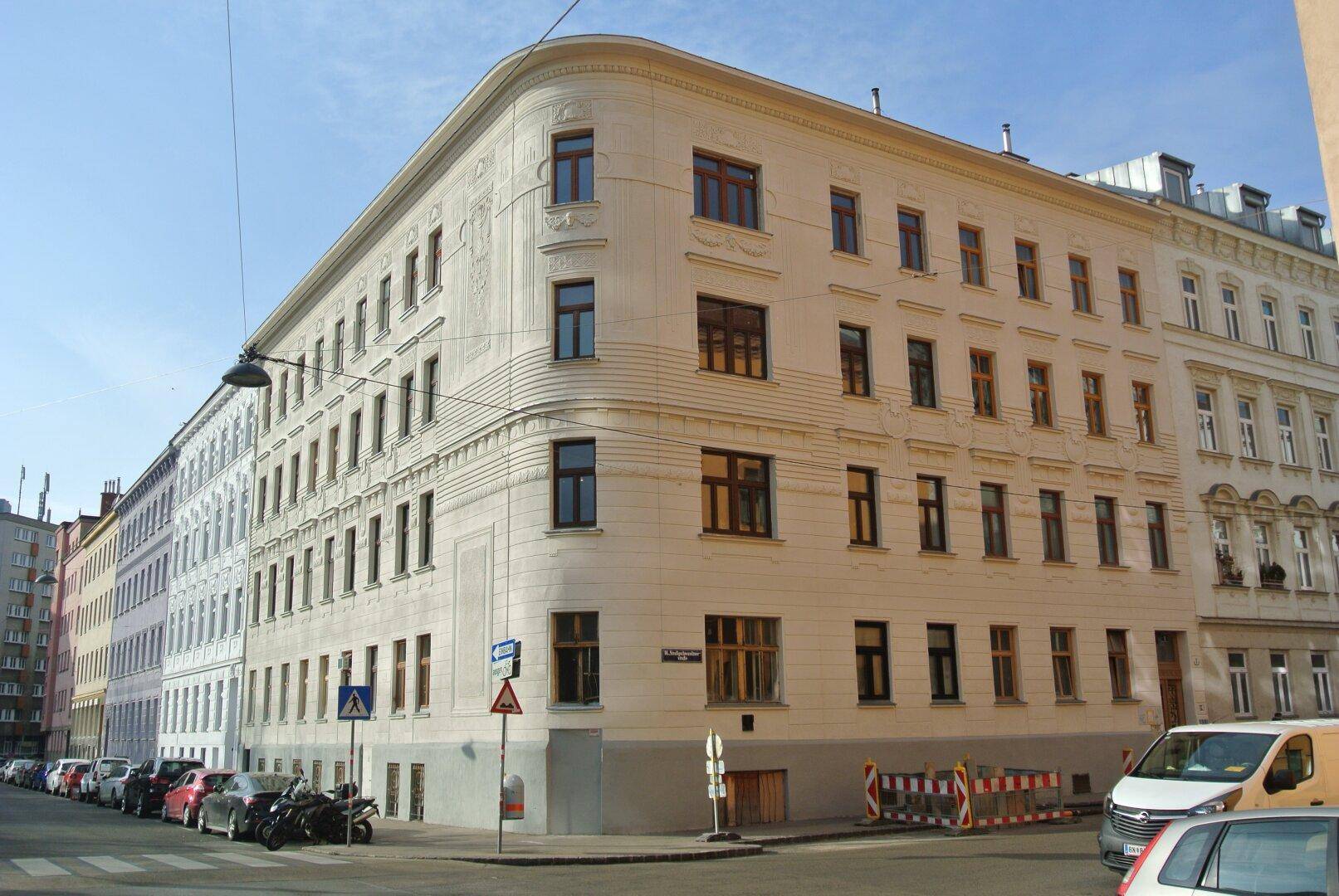 Eckgebäude mit klassischer Architektur und zahlreichen Fenstern, umgeben von einer städtischen Straße mit parkenden Autos.