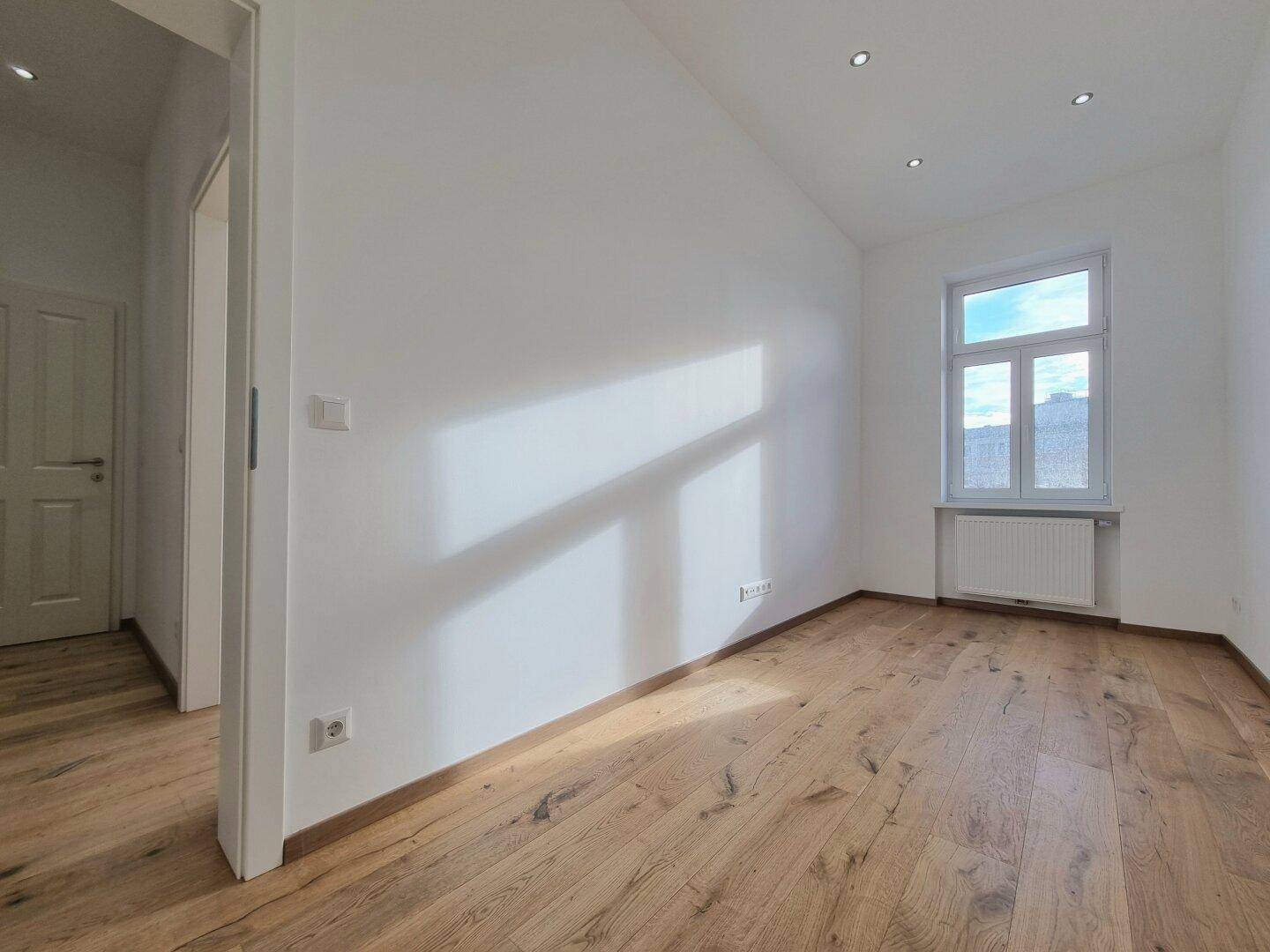 Helles Zimmer mit Eichenparkett und moderner Deckenbeleuchtung, ein Fenster lässt viel Sonnenlicht herein.