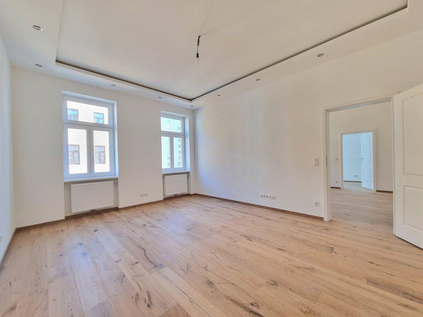 Heller, leerer Raum mit modernem Parkettboden und zwei großen Fenstern.