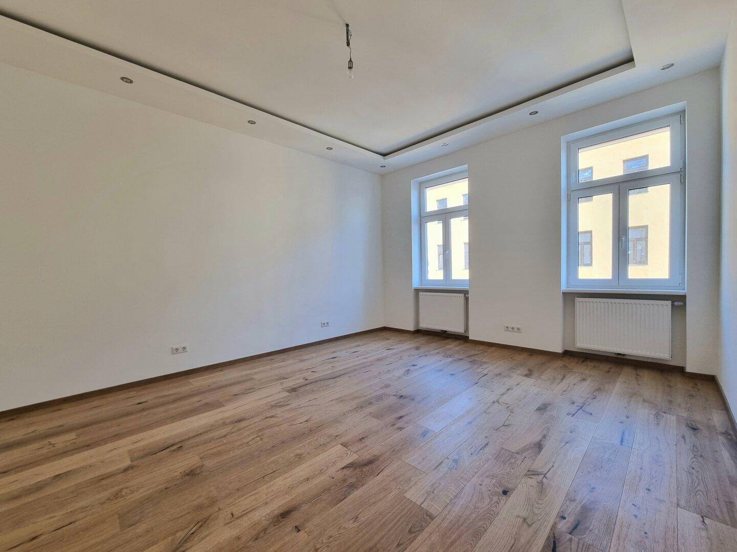 Heller, geräumiger Raum mit Holzboden, weißen Wänden und zwei großen Fenstern.