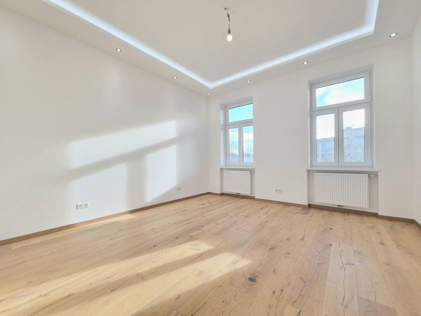 Lichtdurchflutetes Zimmer mit Holzboden, zwei Fenstern und moderner Deckenbeleuchtung.