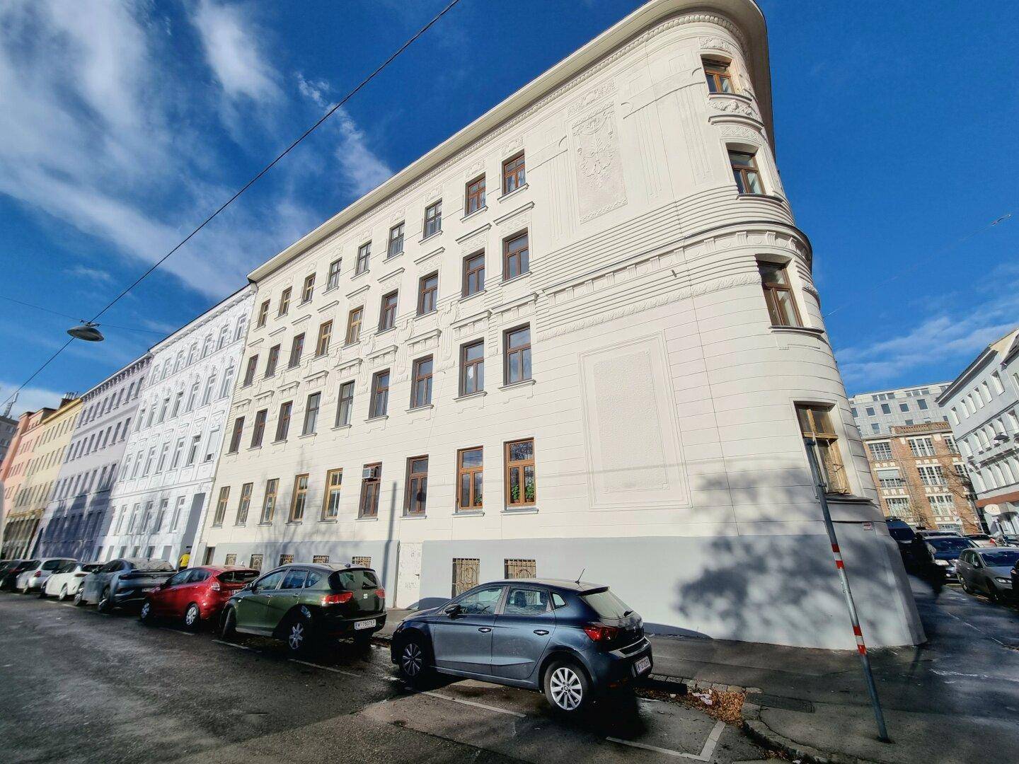 Weitwinkelaufnahme des historischen Eckgebäudes mit langer Straßenansicht und parkenden Fahrzeugen.