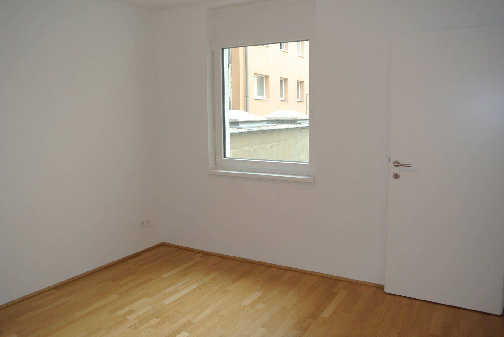 Helles, leeres Zimmer mit weißen Wänden, einem Fenster und hellem Holzboden.