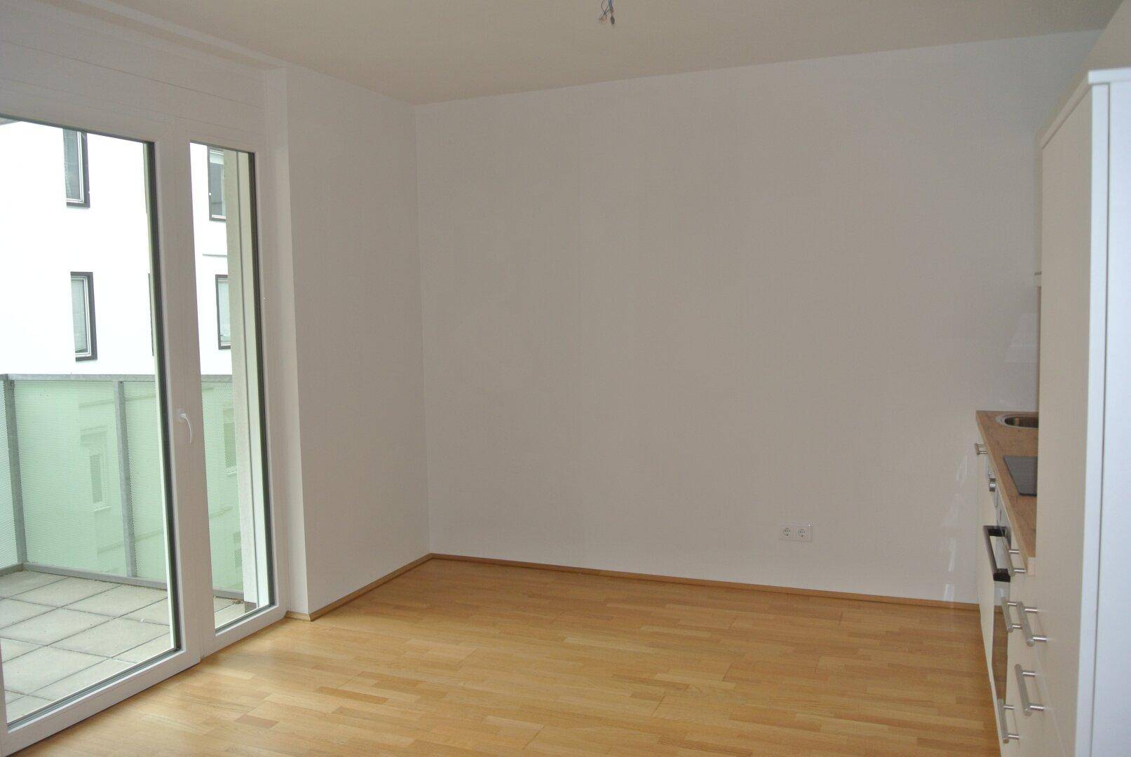 Heller Wohnbereich mit Holzboden, Balkonzugang und integrierter Küchenzeile, bereit zur Gestaltung.