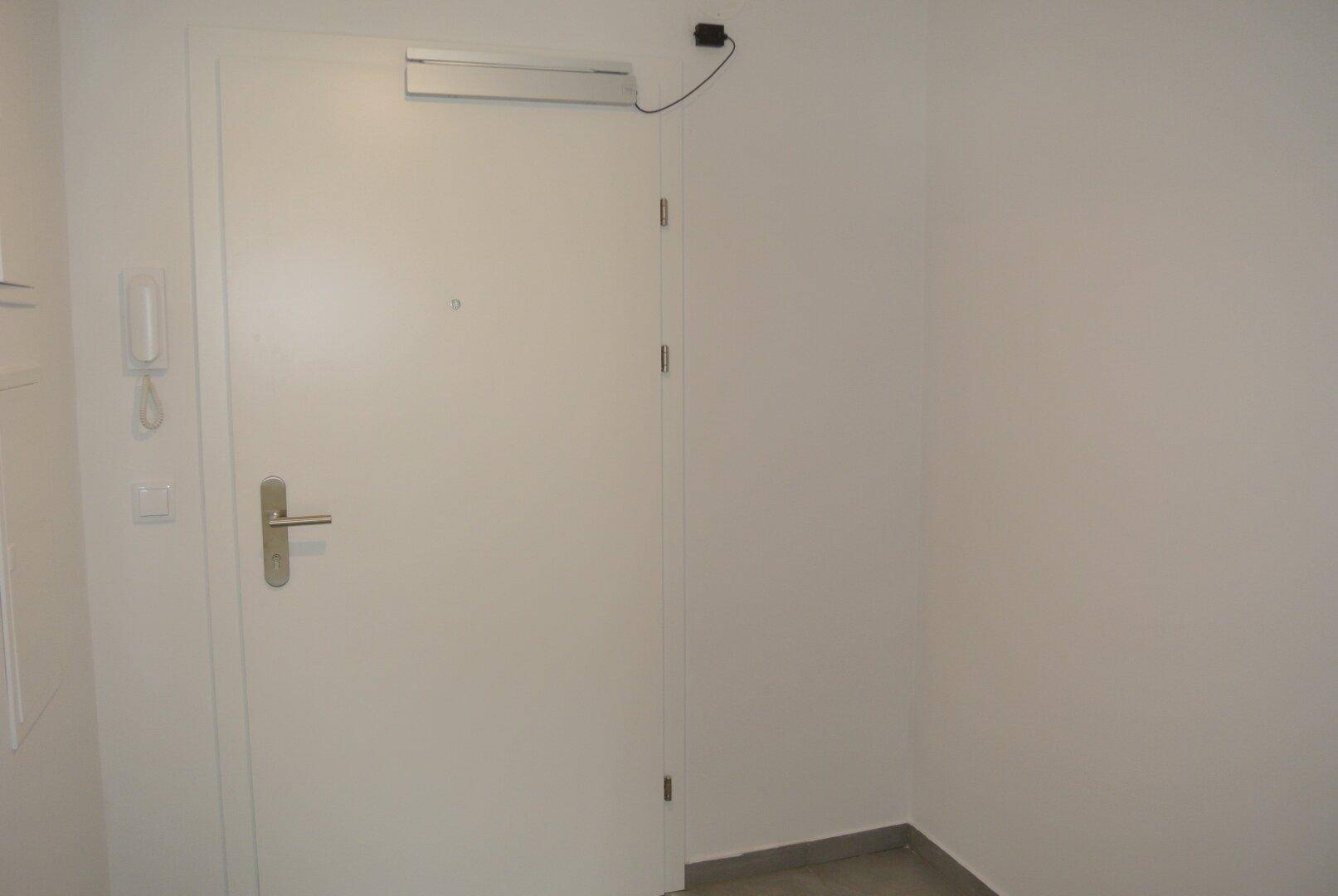 Heller Eingangsbereich einer Wohnung mit weißer Tür, Gegensprechanlage und schlichten Wänden.