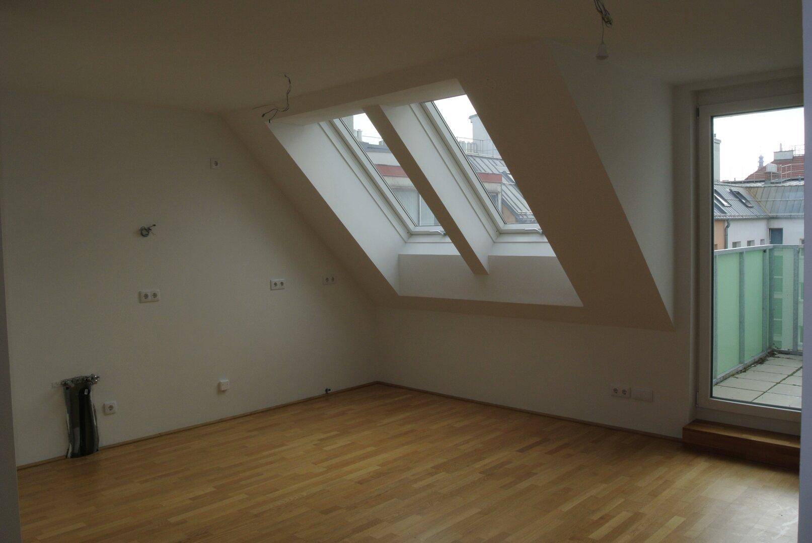 Heller Dachgeschossraum mit Holzfußboden, mehreren Dachfenstern und Balkonzugang.