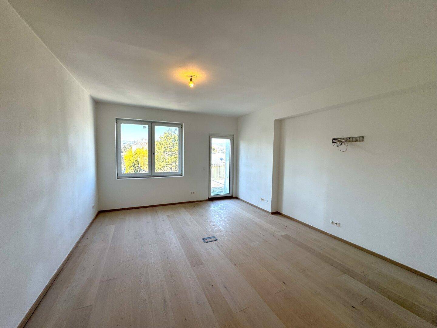 Helles, leeres Zimmer mit hellem Holzboden, weißen Wänden und Zugang zu einem kleinen Balkon.
