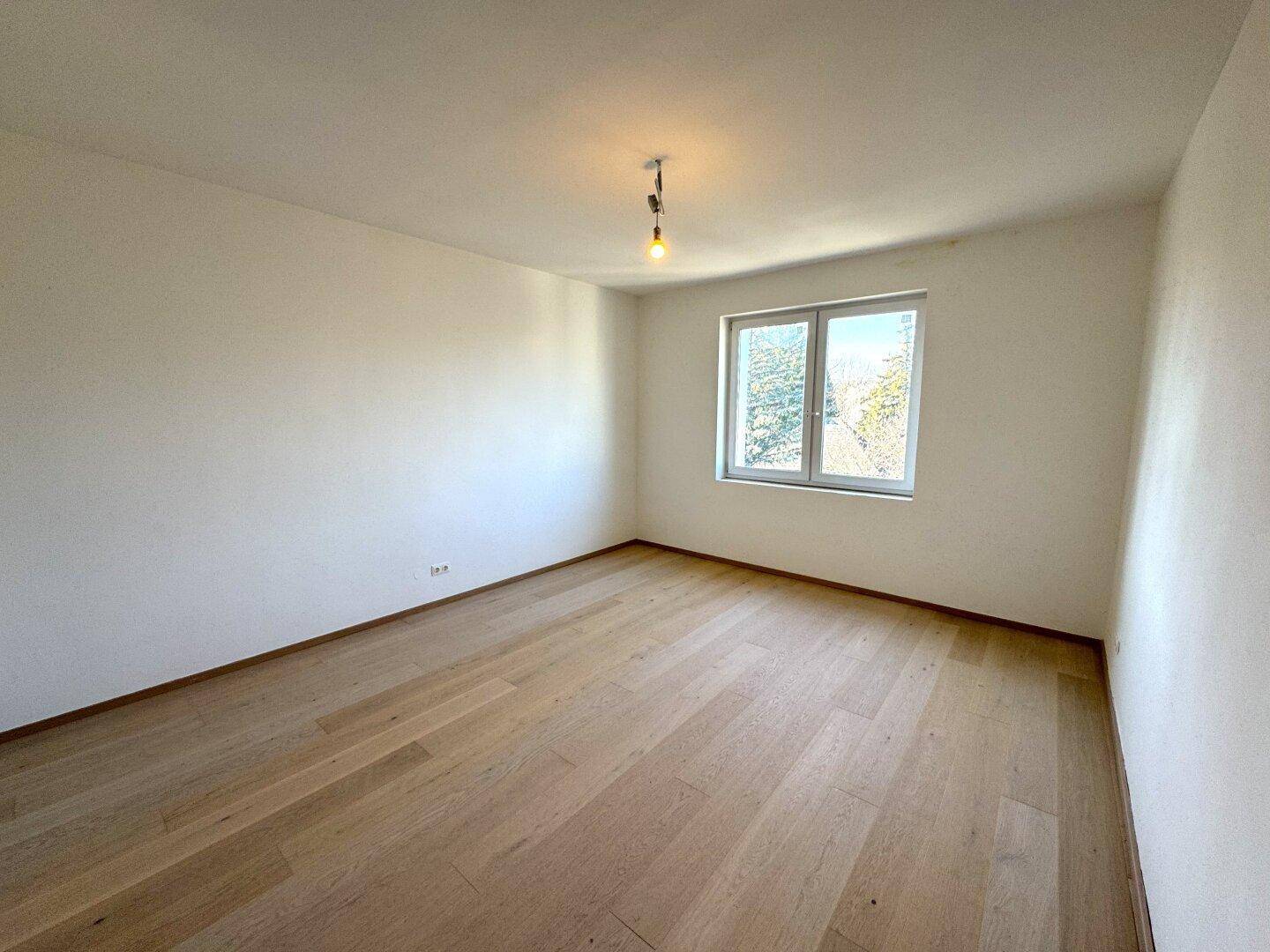 Leeres Schlafzimmer mit hellem Holzboden, weißen Wänden und einem großen Fenster für viel Tageslicht.