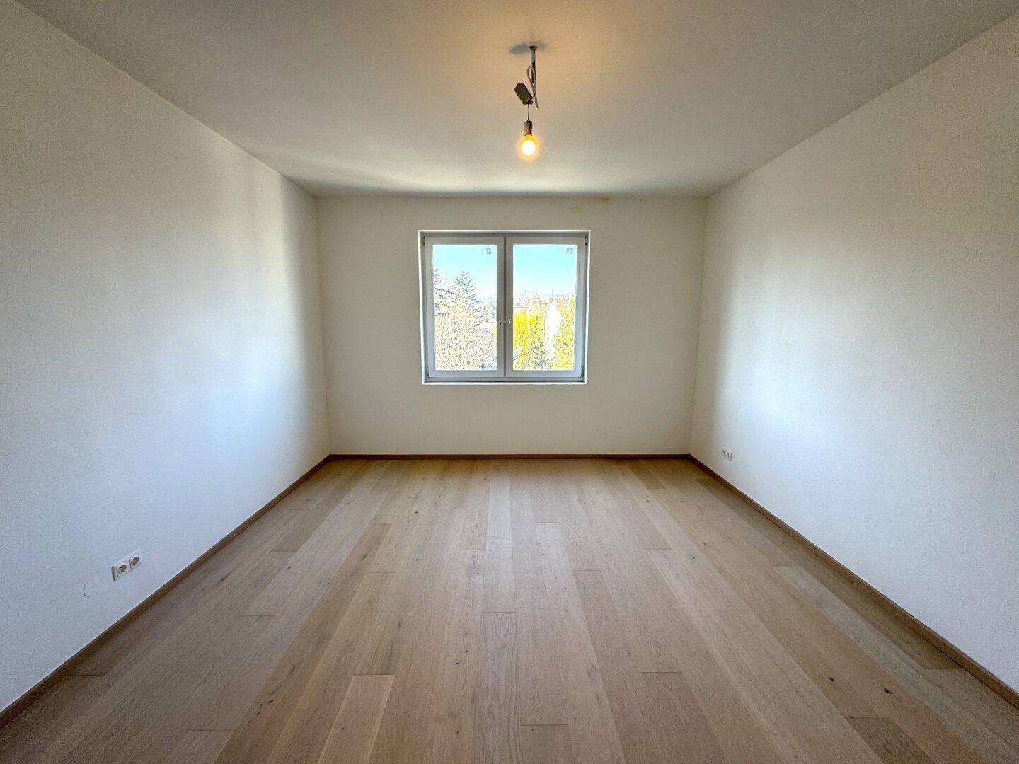 Geräumiges, leeres Zimmer mit hellem Holzboden und einem großen Fenster, das viel Licht hereinlässt.