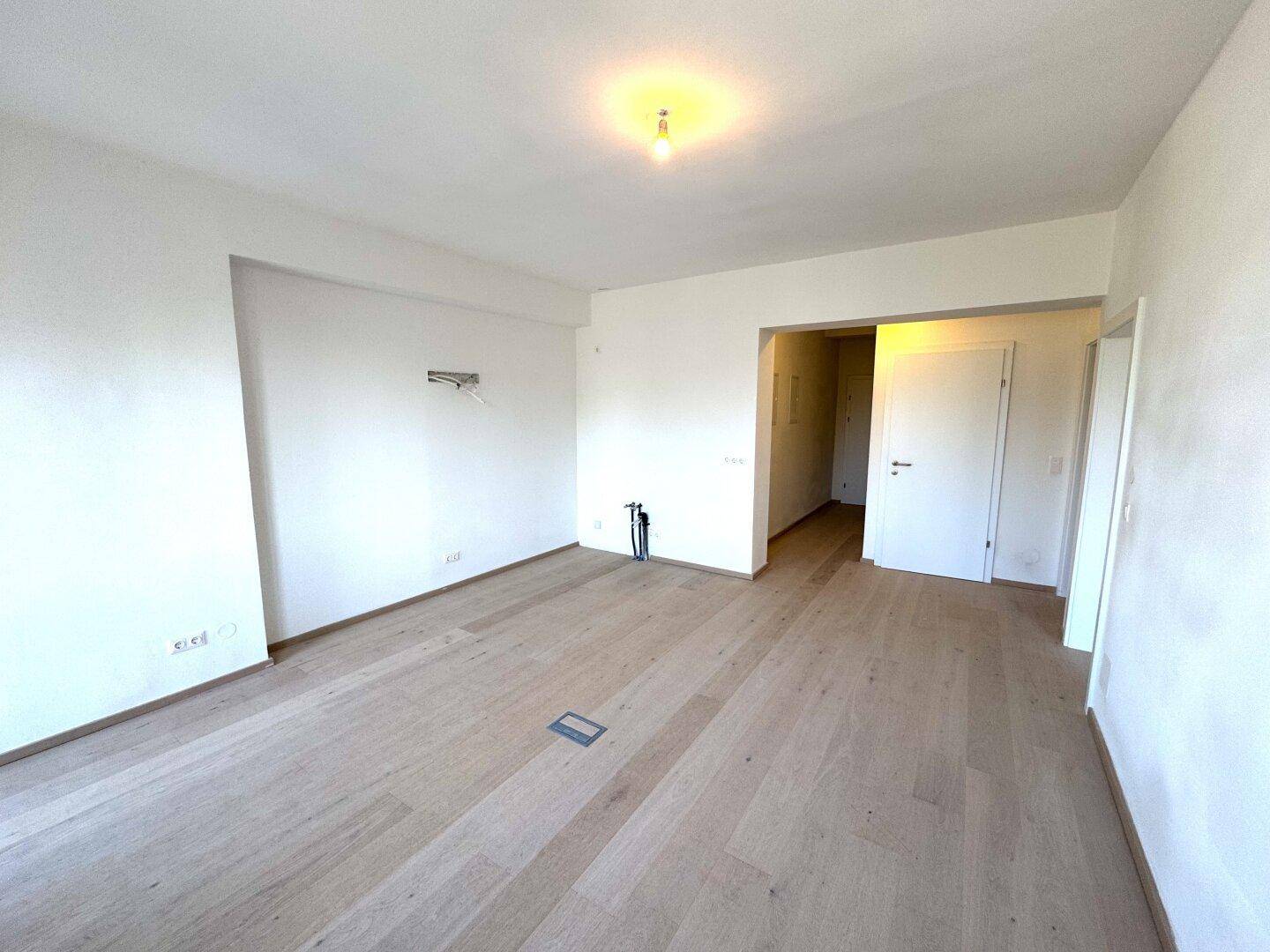 Heller, leerer Raum mit Holzboden, weißen Wänden und offenem Zugang zu weiteren Bereichen.
