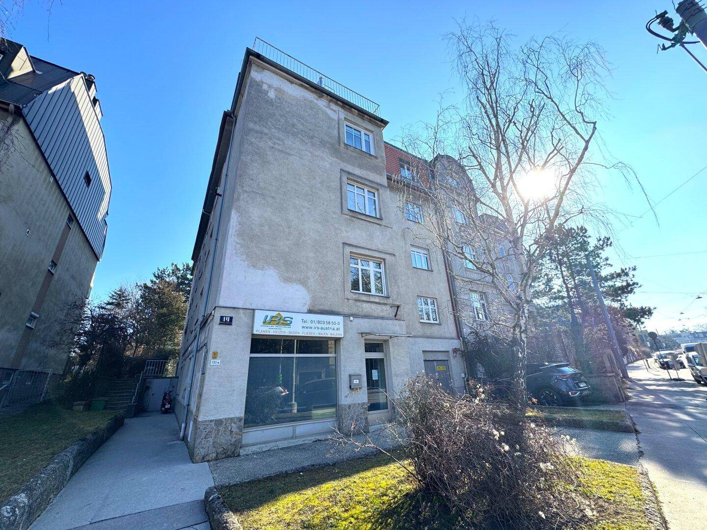 Älteres Gebäude mit Geschäftsfläche im Erdgeschoss und sonniger Fassade unter blauem Himmel.