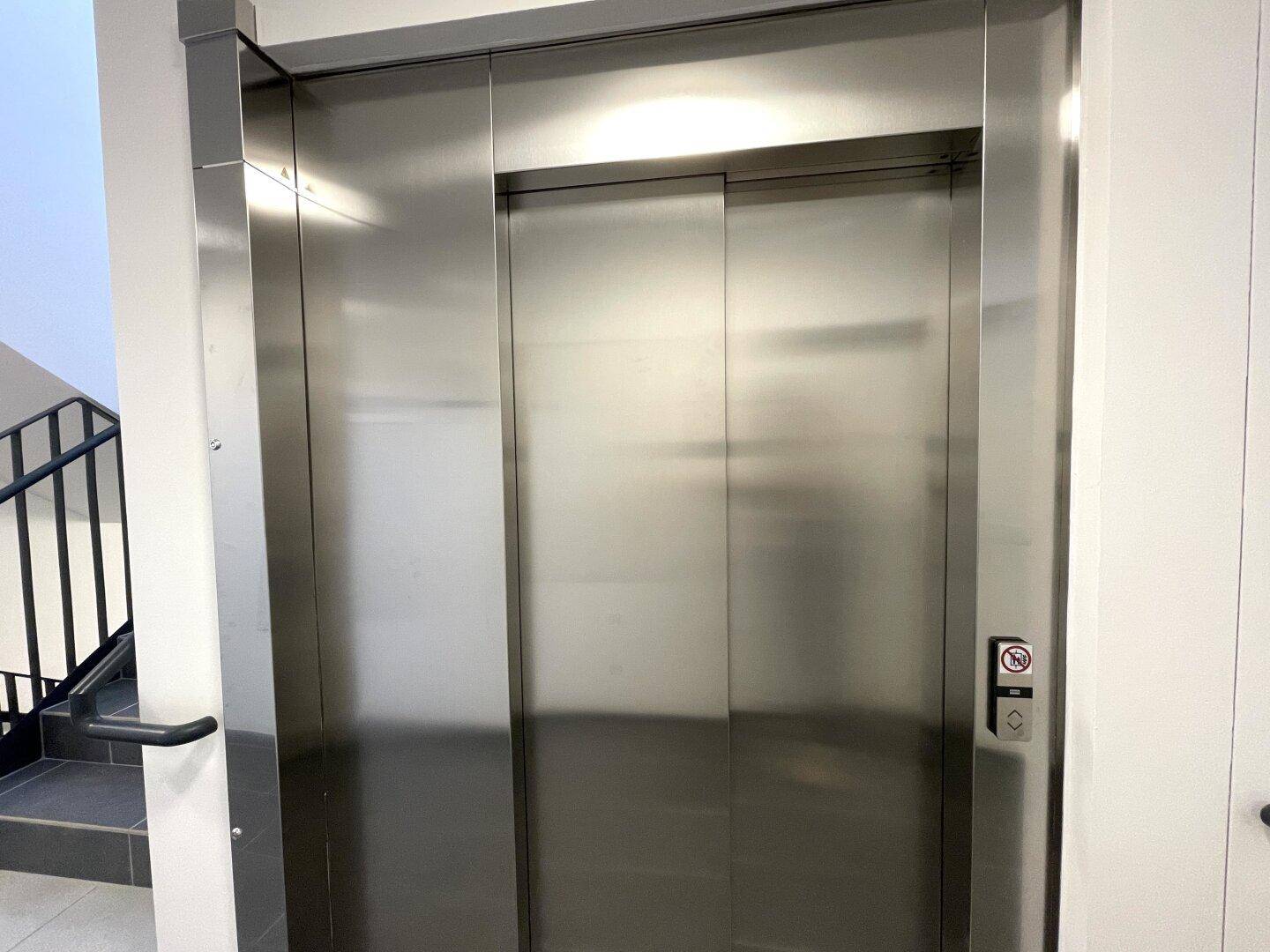 Moderner Aufzug mit Edelstahltüren in einem hellen Treppenhausbereich.