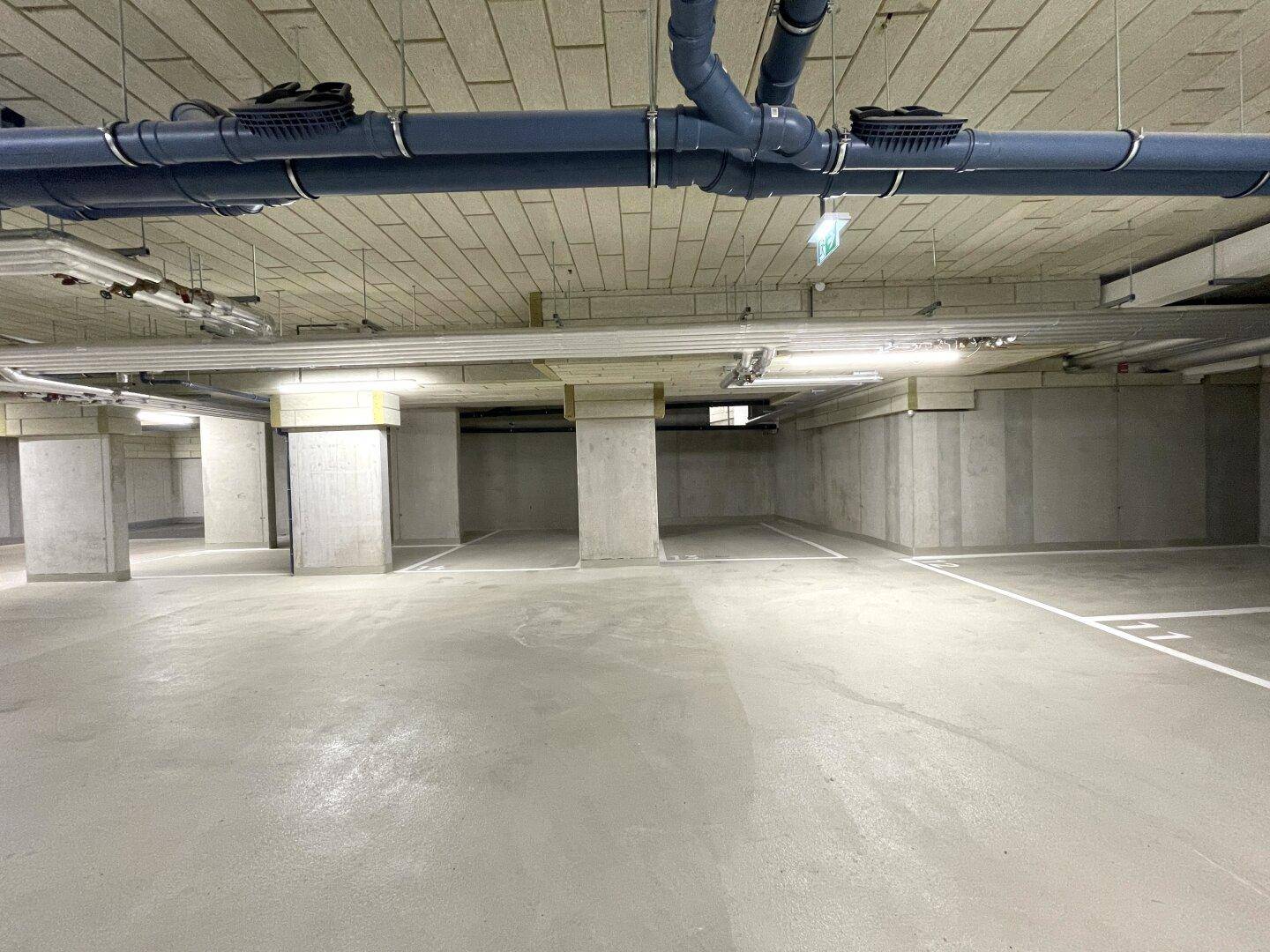 Geräumige Tiefgarage mit Betonstützen, sichtbaren Leitungen und markierten Parkplätzen.
