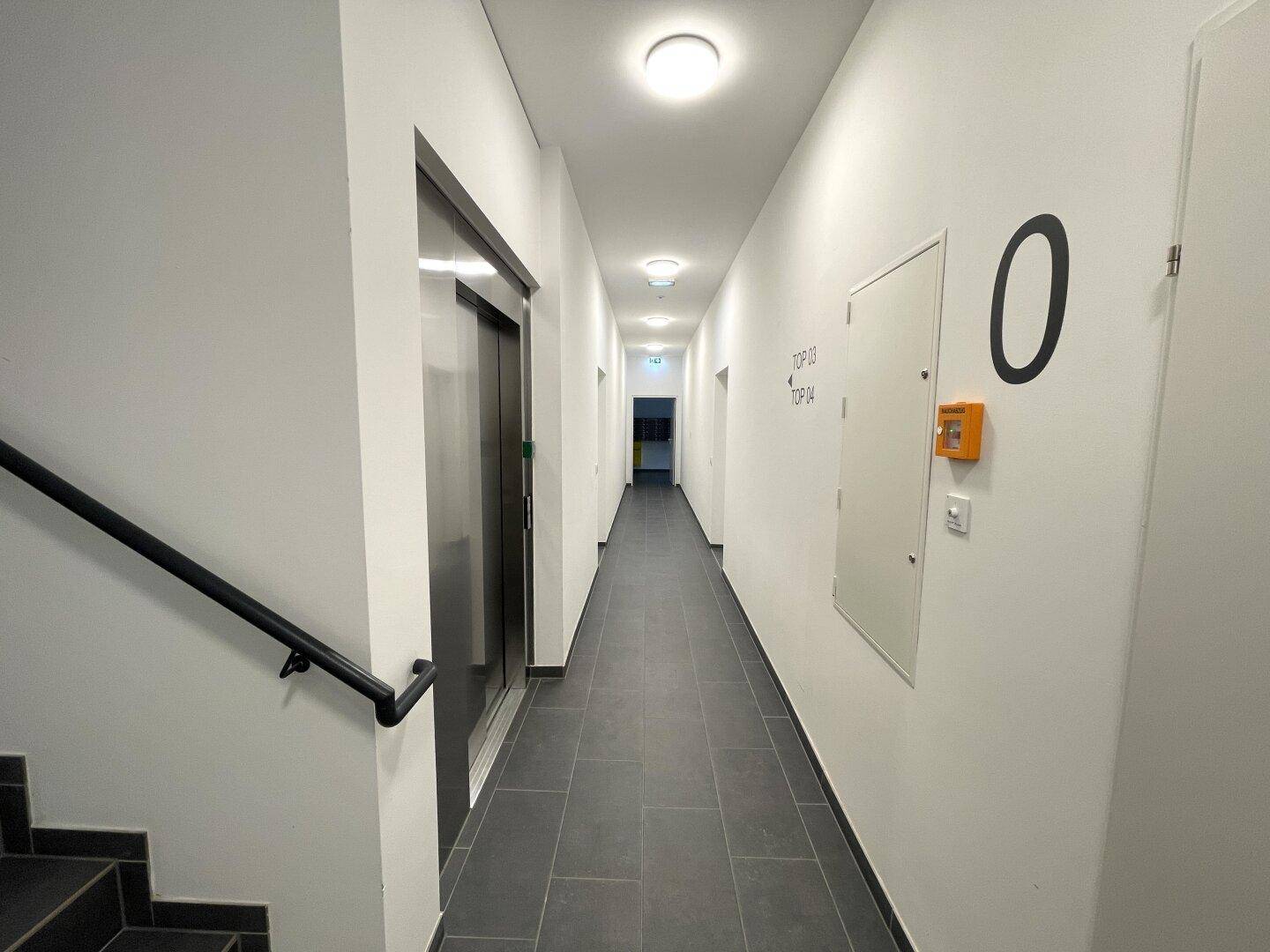 Heller Flur mit Fliesenboden, weiß gestrichenen Wänden und Zugang zum Aufzug.