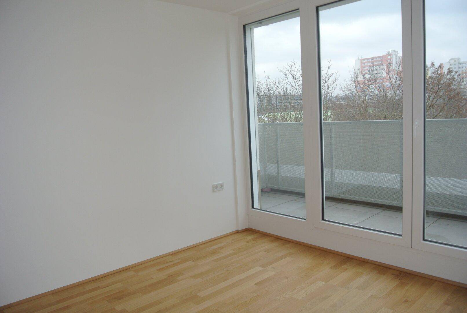 Lichtdurchfluteter Raum mit Holzboden und breiter Fensterfront zum Balkon.