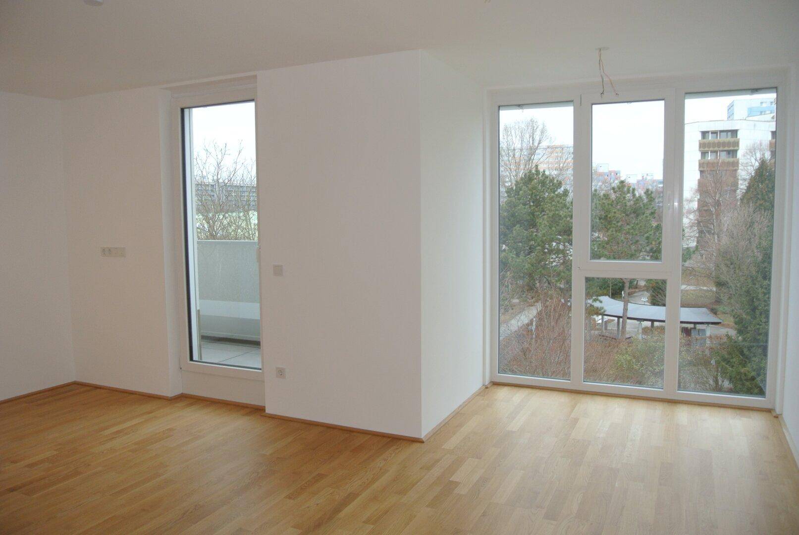Helles, unmöbliertes Wohnzimmer mit großen Fenstern, Holzboden und Zugang zu einem Balkon.