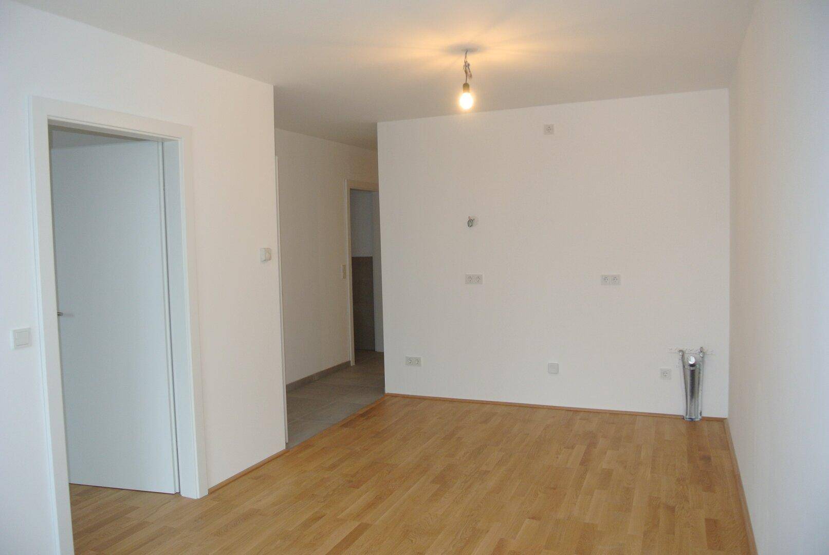 Heller, leerer Raum mit Holzboden und weiß gestrichenen Wänden, der zu weiteren Zimmern führt.