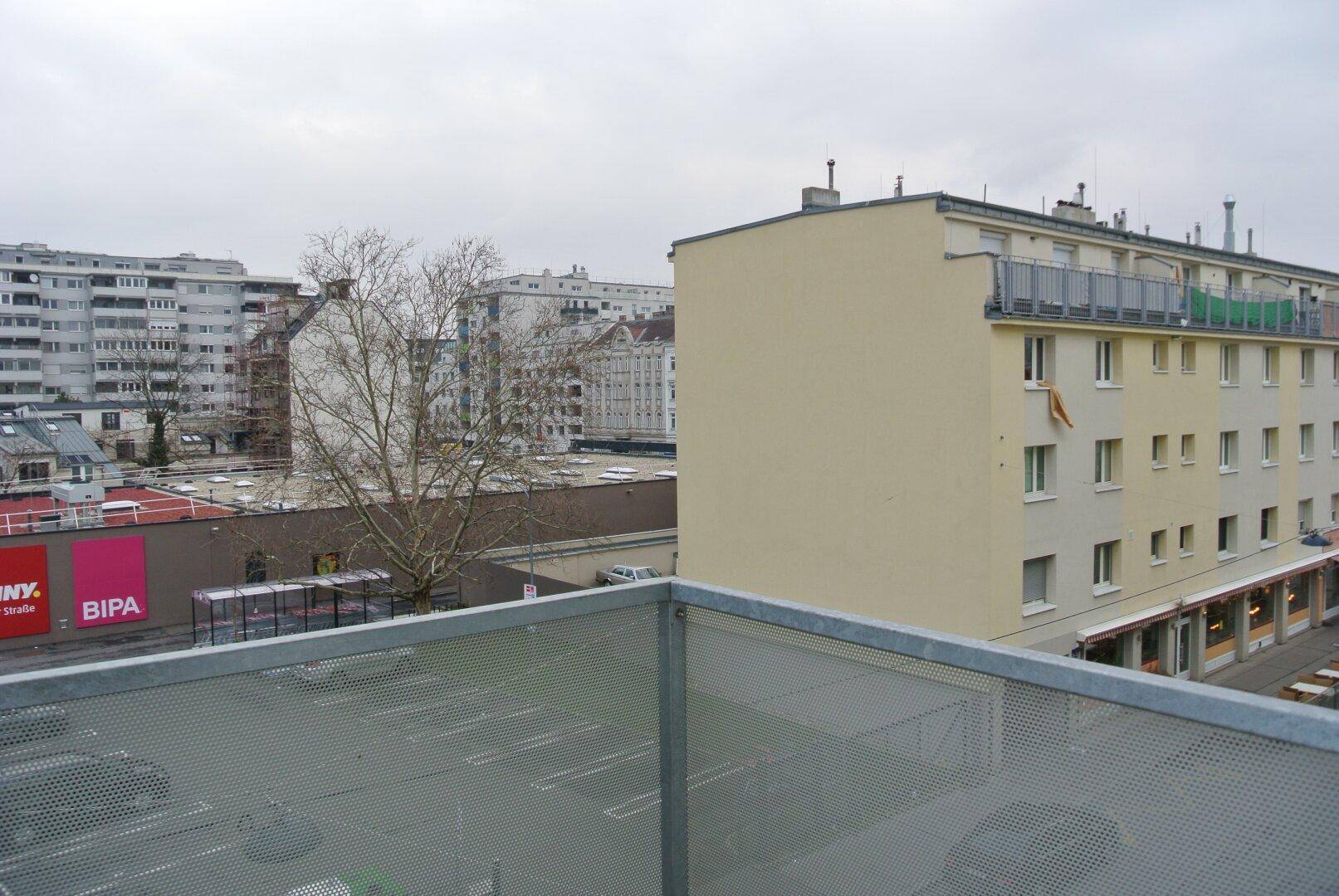 Blick von einem Balkon auf die umliegende Stadtbebauung mit Geschäften und Parkplätzen.