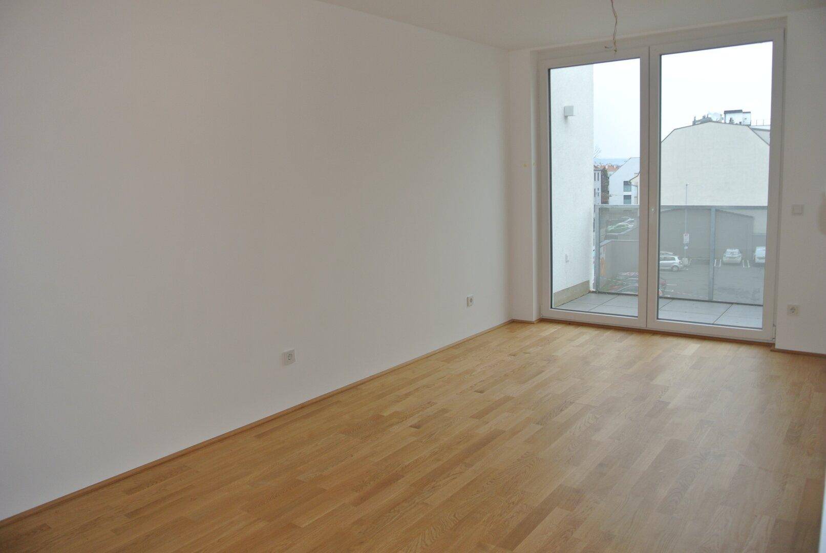 Lichtdurchfluteter Wohnraum mit Holzboden und großem Fenster, das Zugang zu einem Balkon bietet.