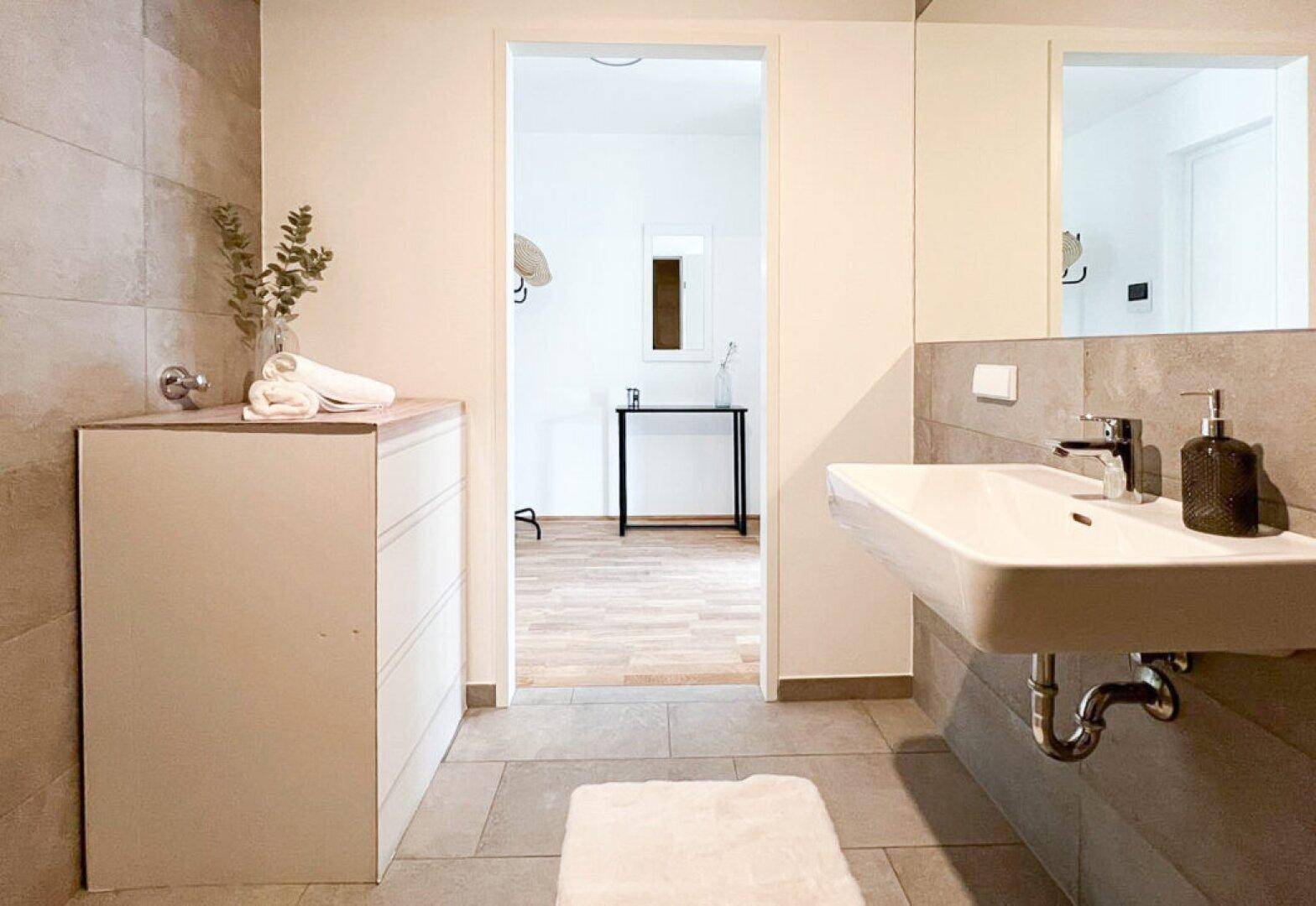 Modernes Badezimmer mit Waschbecken, Spiegel und hellen Fliesen, angrenzend an einen WC-Bereich.