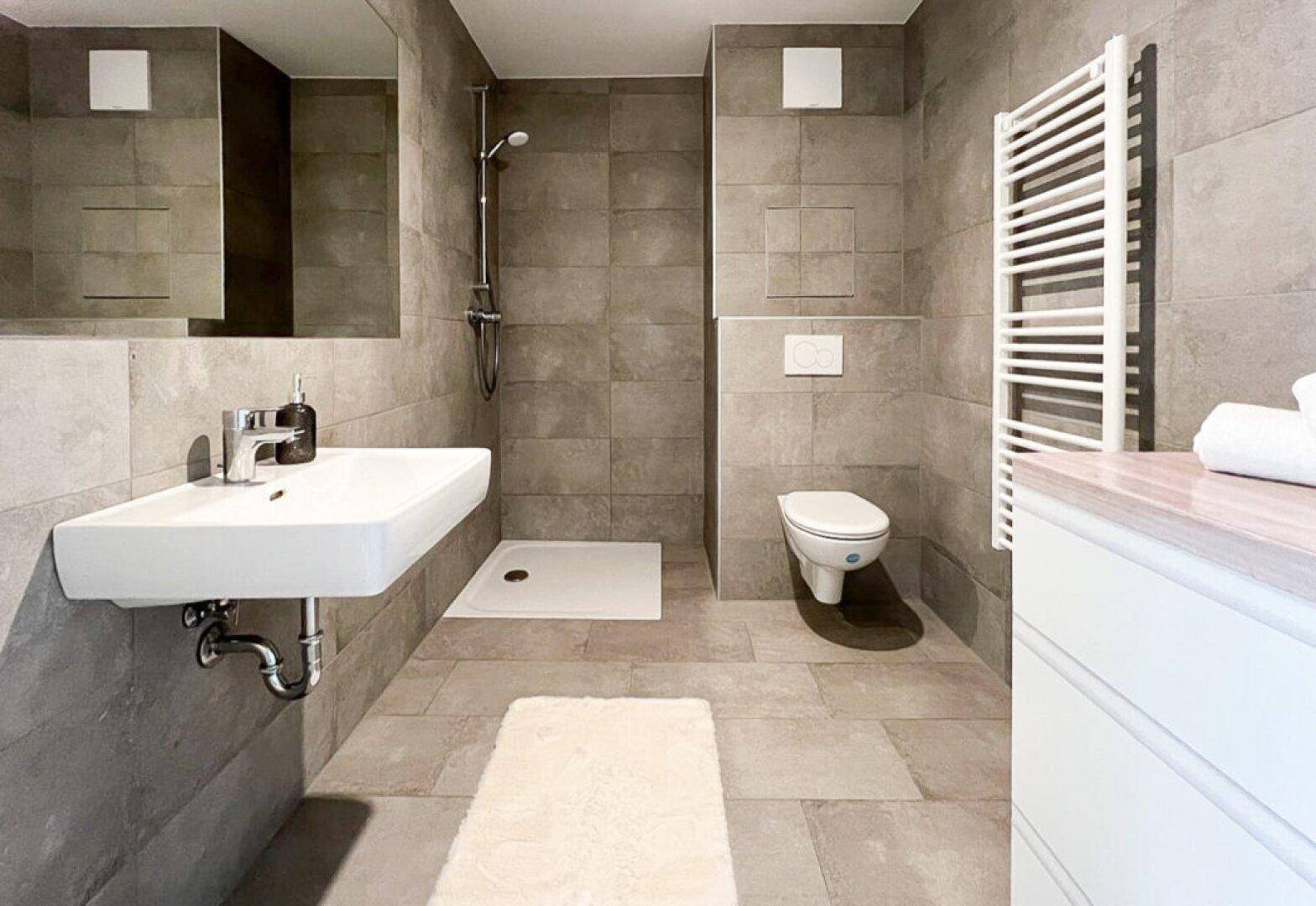 Modernes Badezimmer mit Dusche, wandhängendem WC und beheiztem Handtuchhalter in Grautönen.