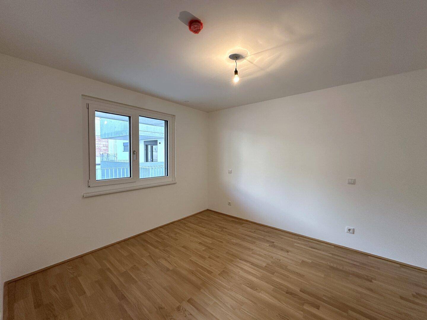 Leeres Schlafzimmer mit hellem Holzboden und großem Fenster für viel Tageslicht.