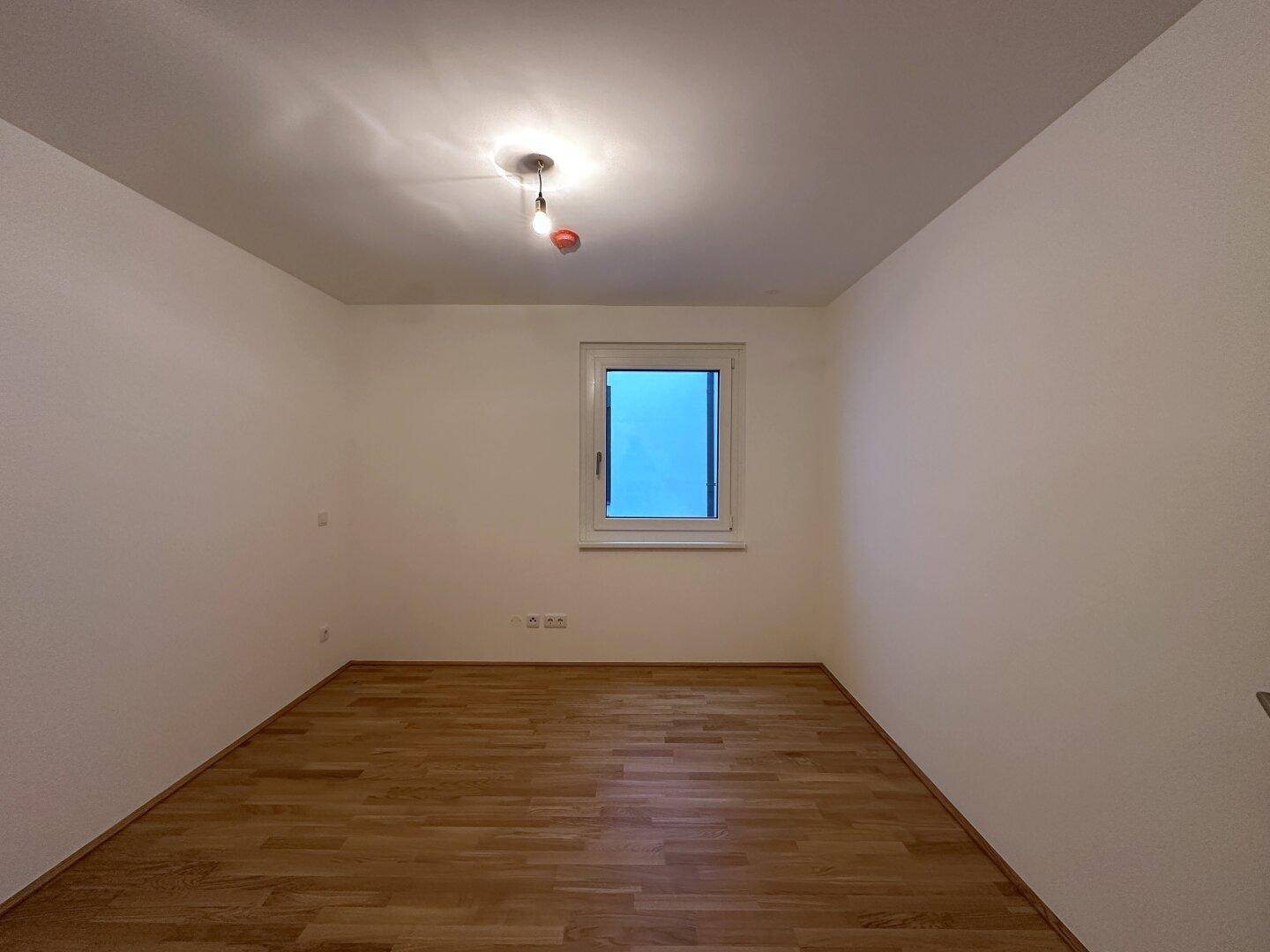 Leeres Zimmer mit Holzboden und Fenster, ideal als Kinder- oder Arbeitszimmer.