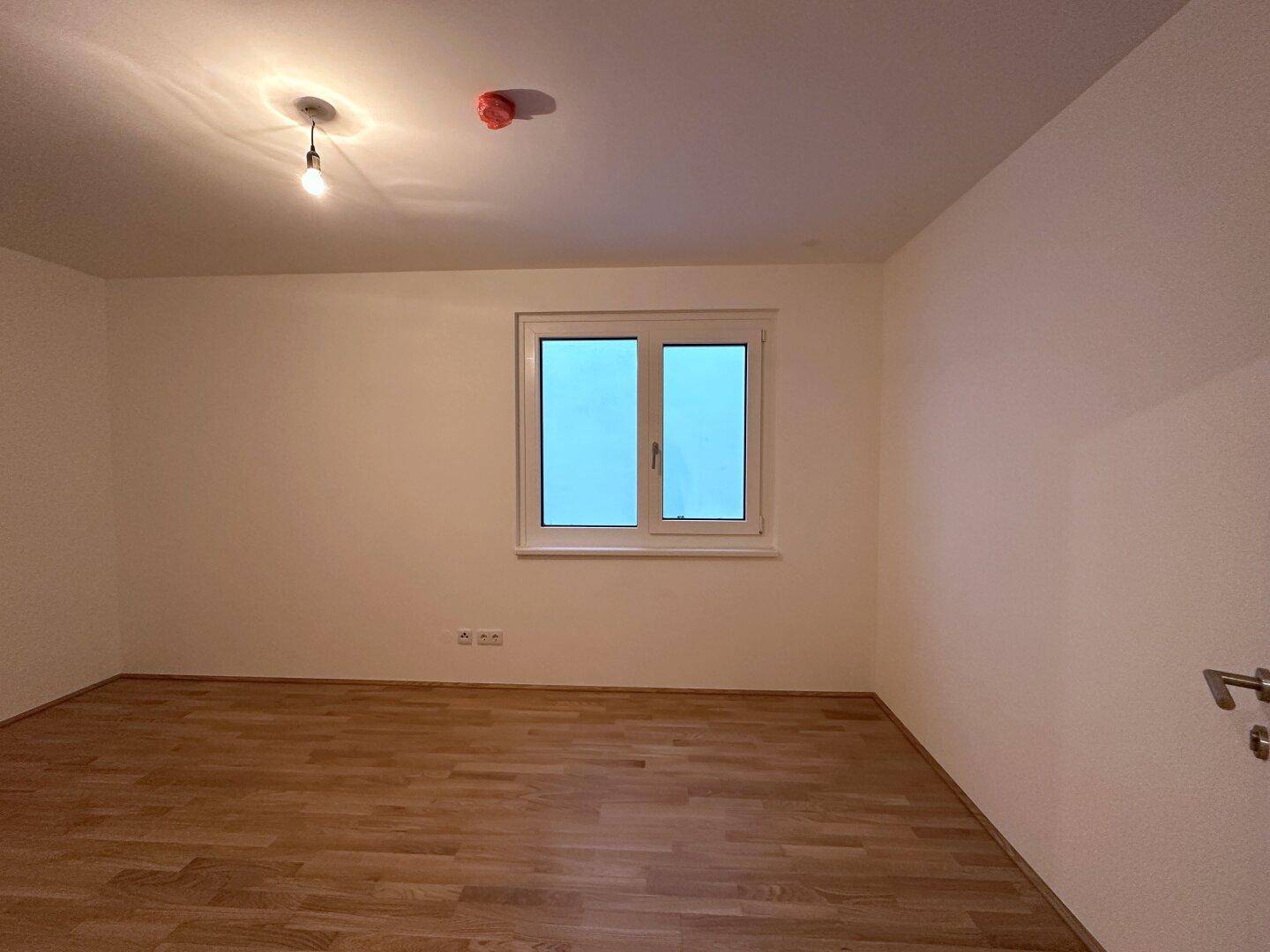 Unmöbliertes Zimmer mit Parkettboden und einem Fenster, das viel Licht hereinlässt.