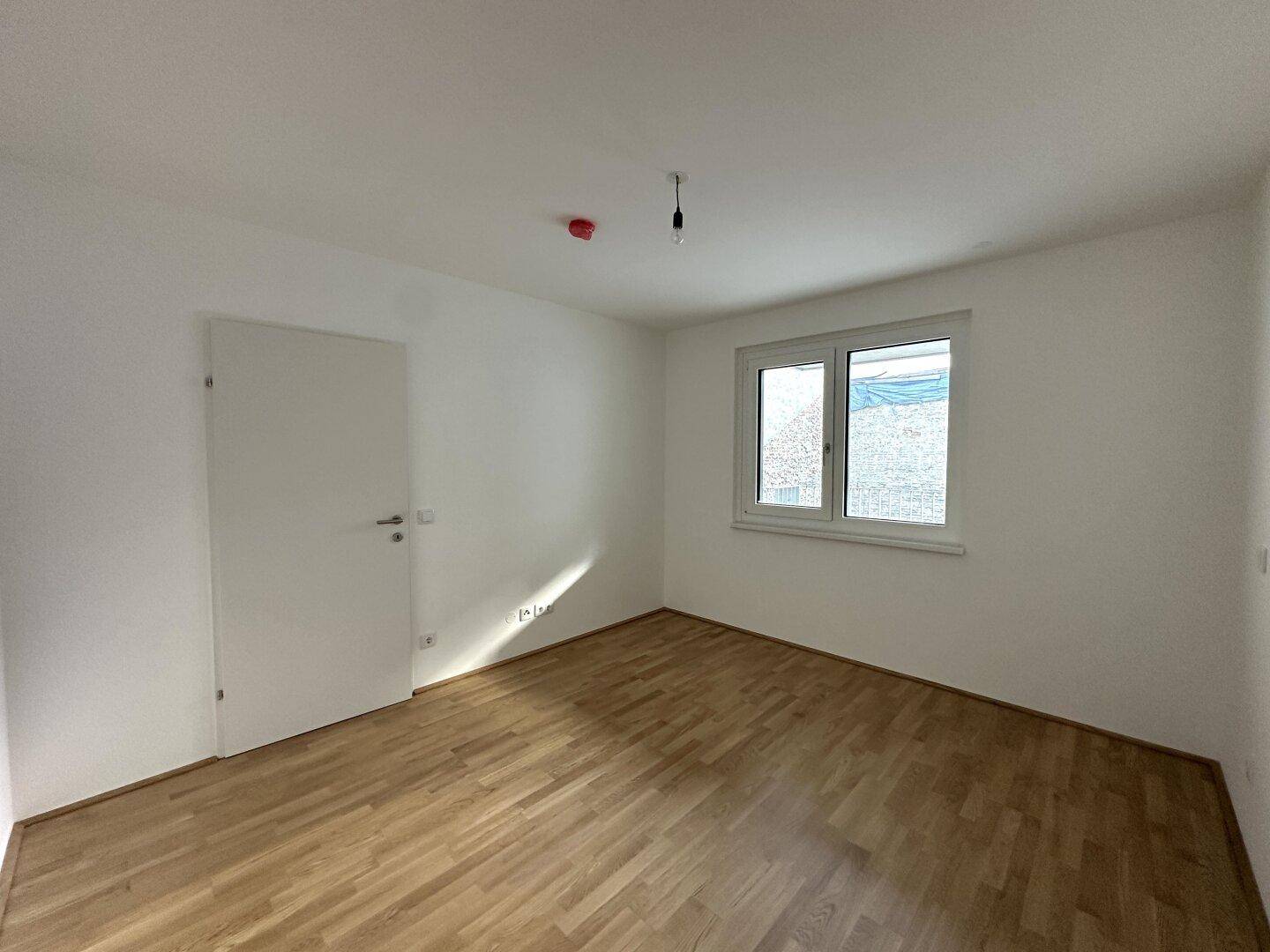 Freundliches Zimmer mit Parkettboden, weißer Tür und Fenster, ideal als Schlafzimmer.