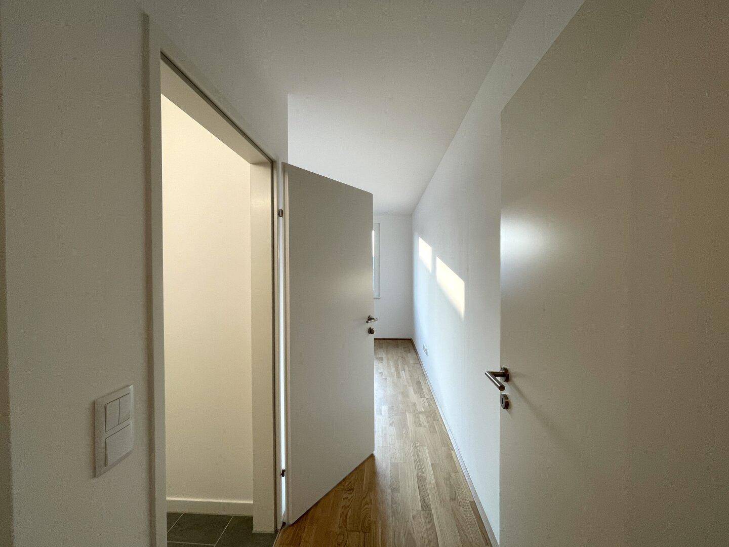 Heller Flur mit Holzboden und offener Tür, die den Zugang zu einem weiteren Zimmer ermöglicht.