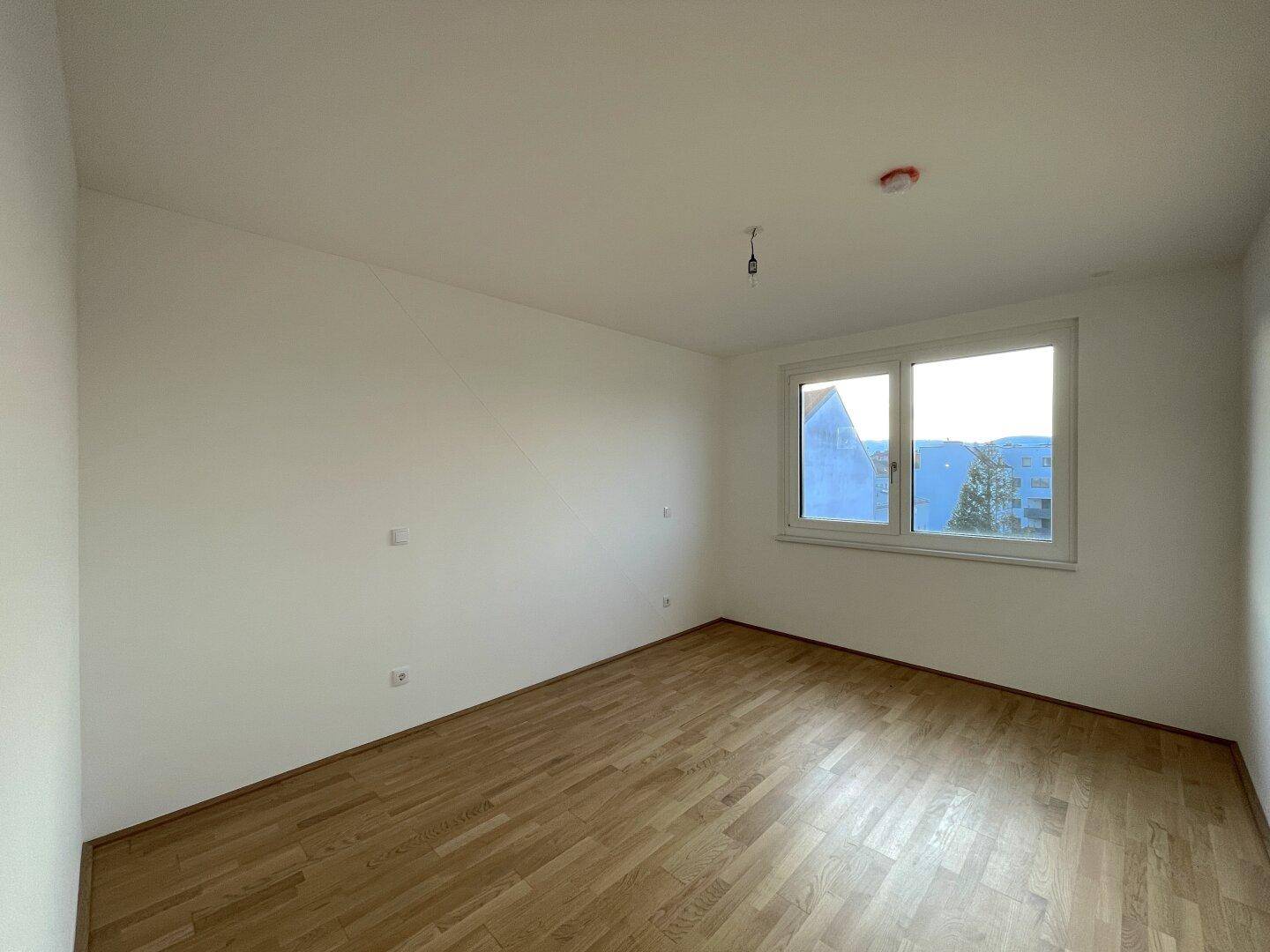 Helles, leeres Zimmer mit Holzboden und einem Fenster, ideal als Schlaf- oder Arbeitszimmer.