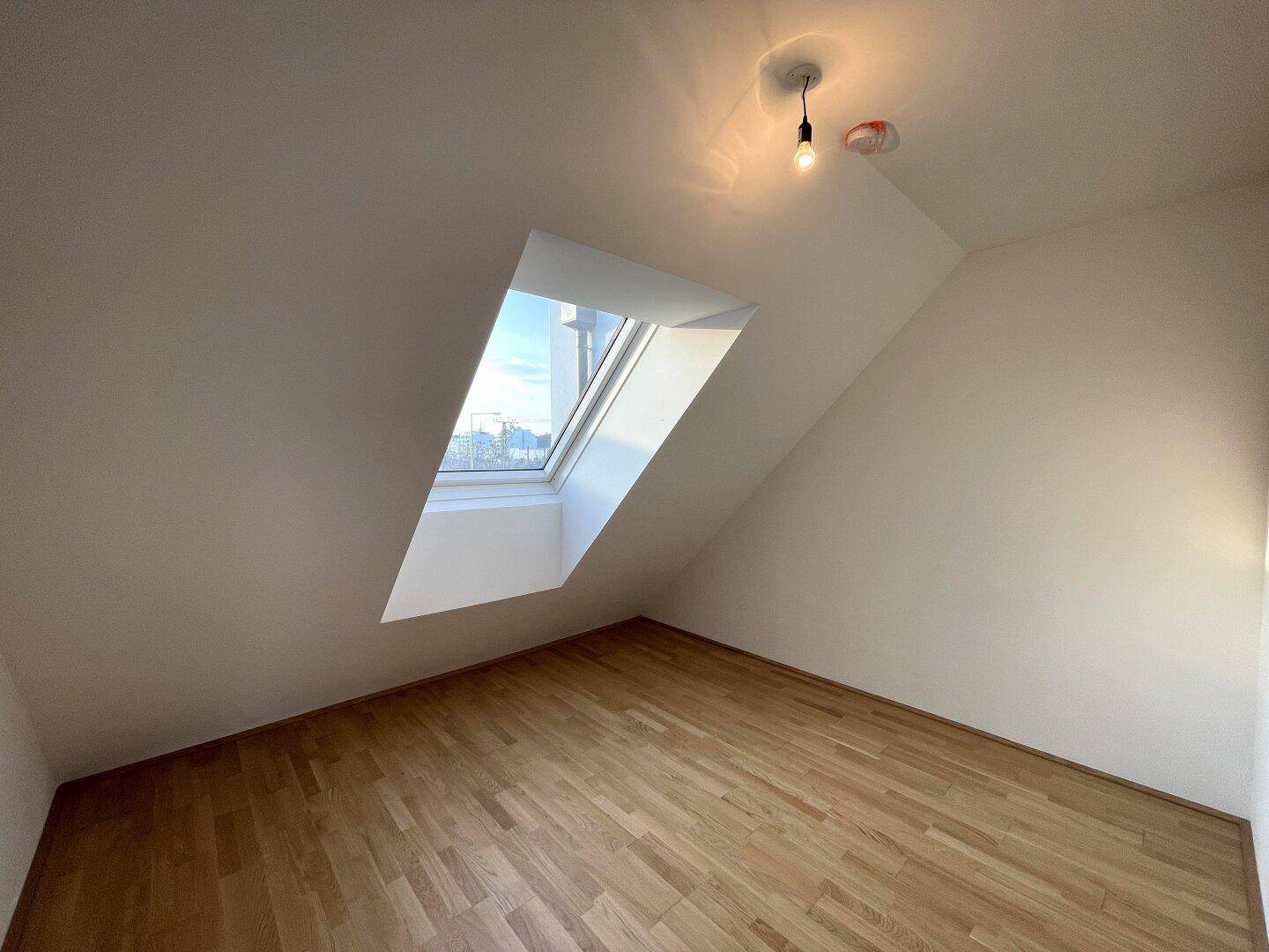 Helles Dachzimmer mit Holzboden und schräger Decke, das durch das Dachfenster viel Licht erhält.