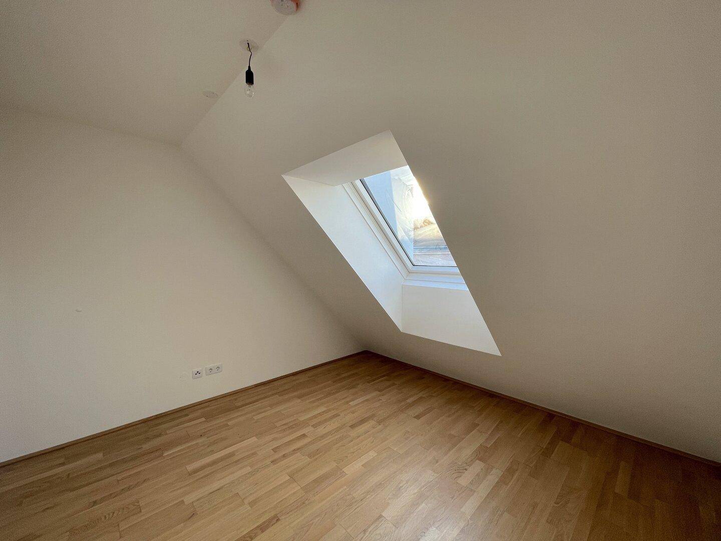 Dachzimmer mit Holzboden und schräger Decke, das durch ein großes Dachfenster hell erleuchtet wird.