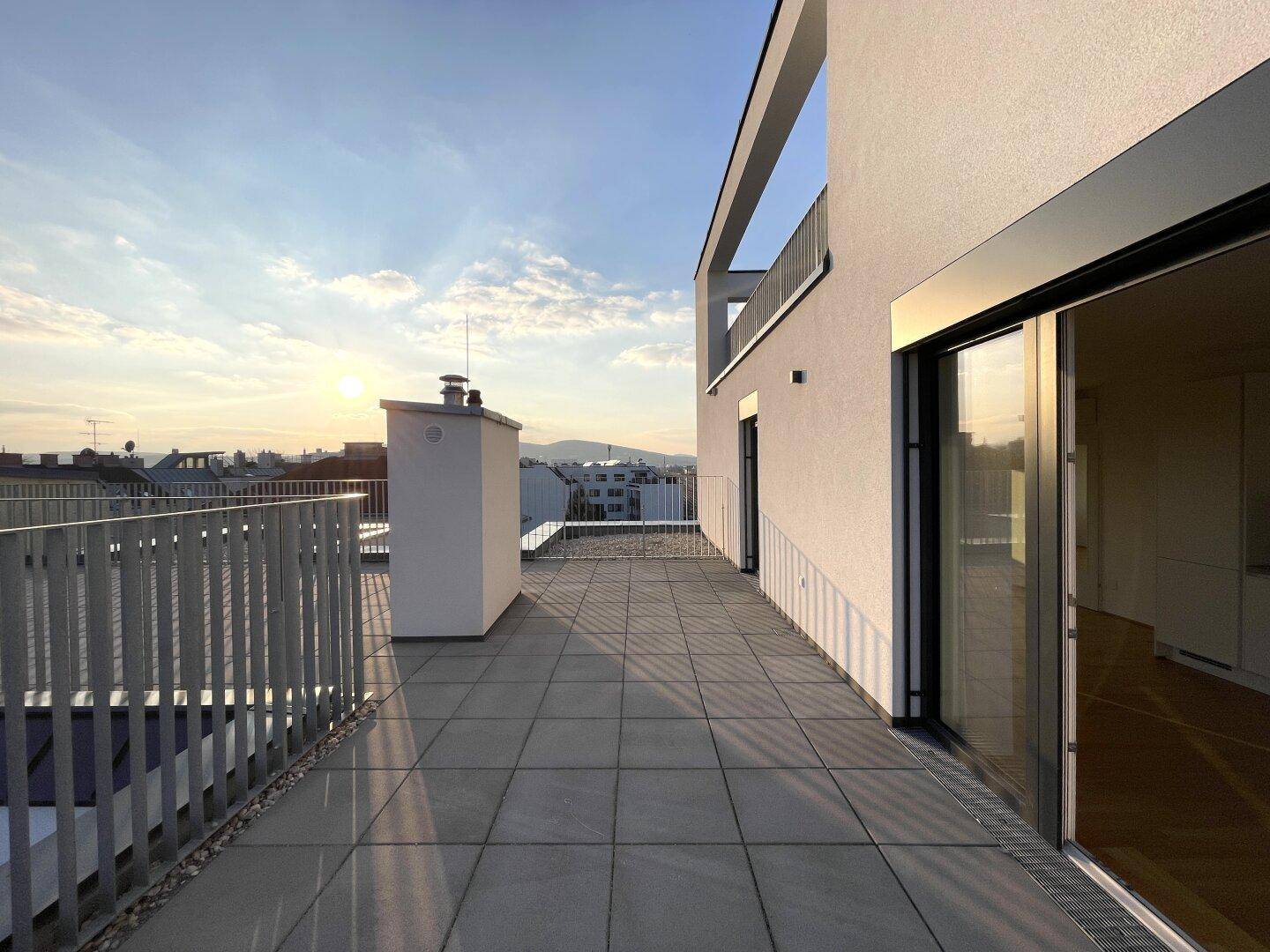 Weitläufige Dachterrasse mit Zugang vom Innenbereich und Blick auf die Stadt bei Sonnenuntergang.