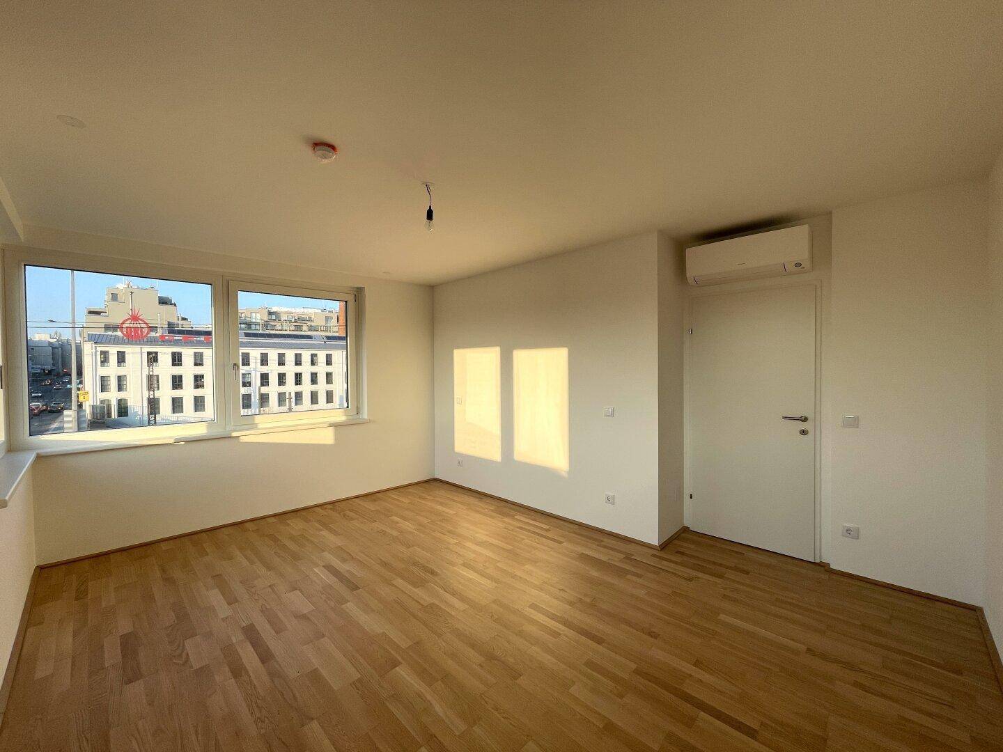 Helles Zimmer mit Parkettboden, Klimaanlage und Fensterblick auf die städtische Umgebung.