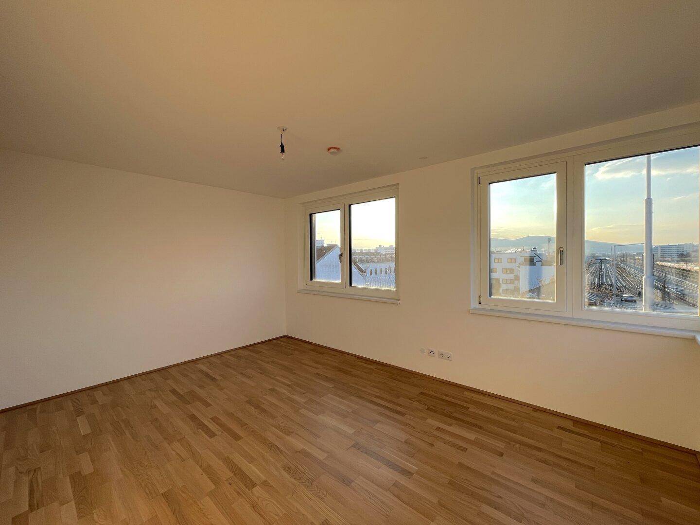 Geräumiges Zimmer mit Parkettboden und Fenstern mit Blick auf die Stadt und Bahnanlagen.
