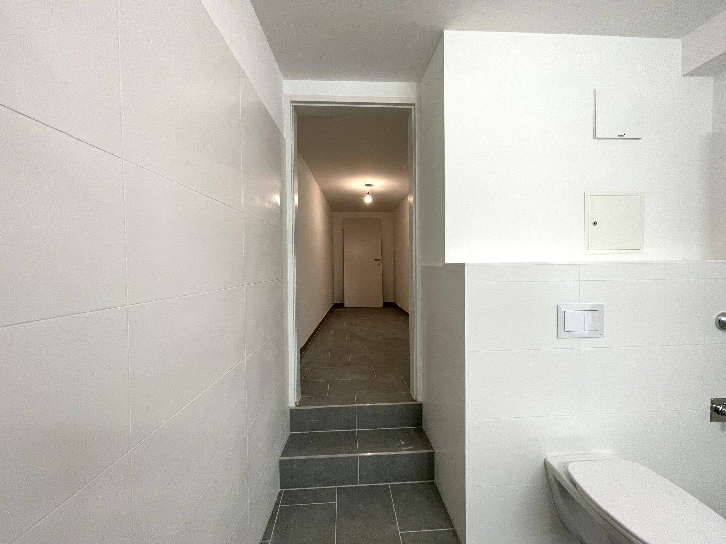 Blick vom Badezimmer in einen gefliesten Flur mit Treppenabsatz und weißer Tür am Ende.