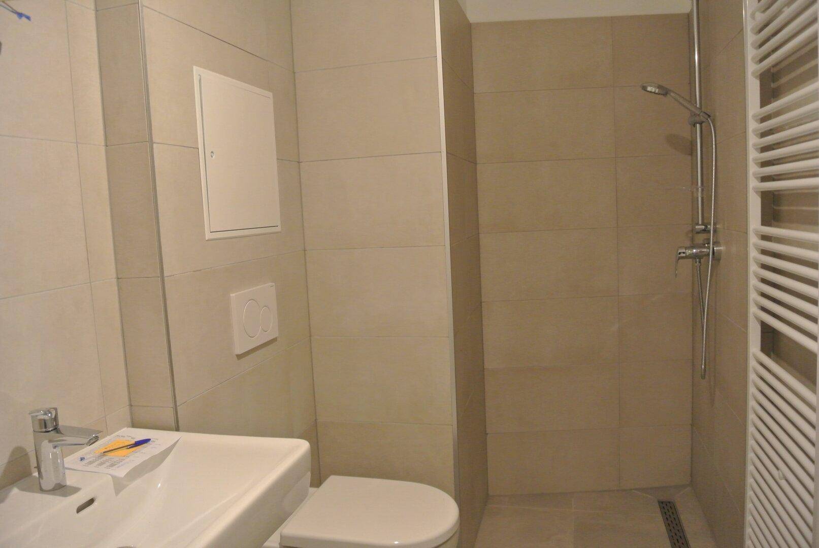 Modernes Badezimmer mit beigefarbenen Fliesen, Dusche, WC und beheiztem Handtuchhalter.