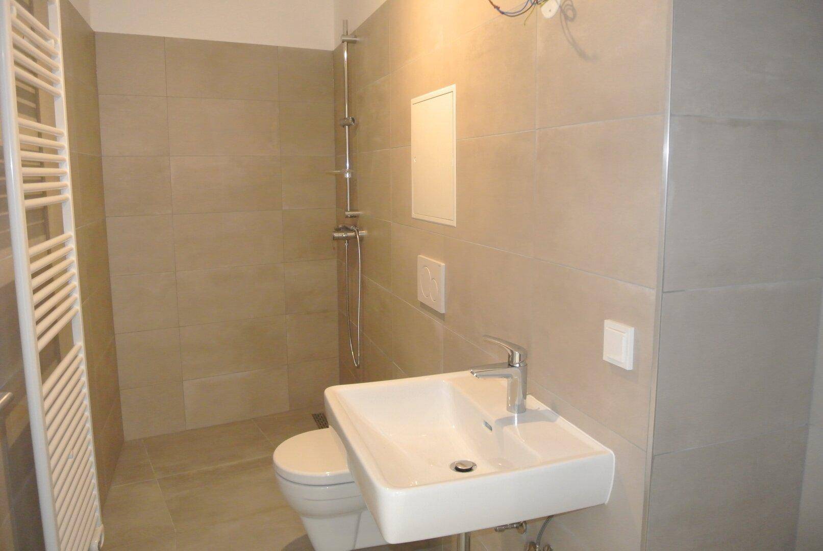 Modernes Badezimmer mit beigefarbenen Fliesen, wandhängendem WC, Dusche und Handtuchheizkörper.