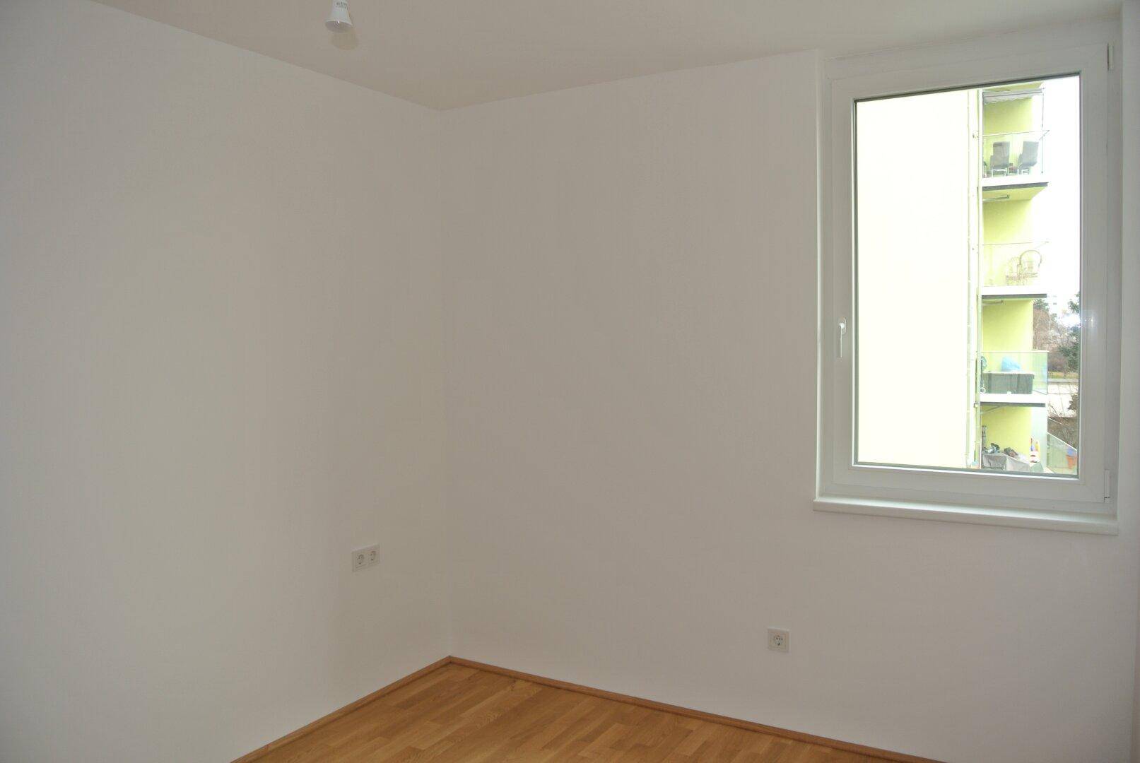 Heller Raum mit Holzboden und einem Fenster, das den Blick auf ein Nachbargebäude freigibt.