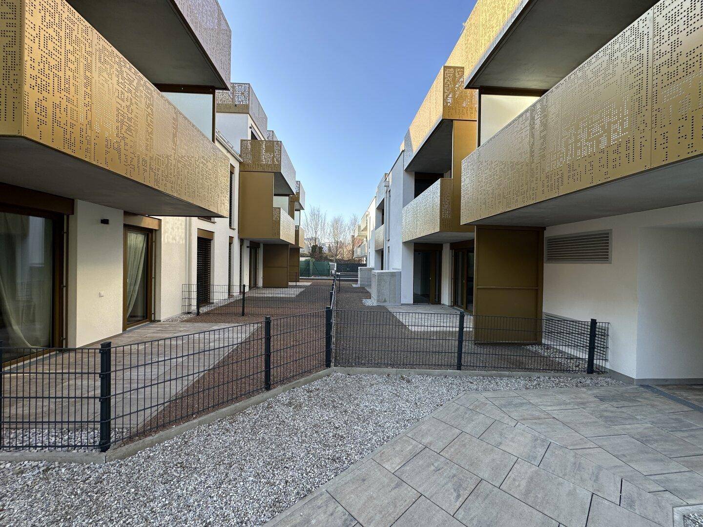 Blick zwischen zwei modernen Gebäudeflügeln mit perforierten goldenen Balkonen und gepflasterten Wegen.