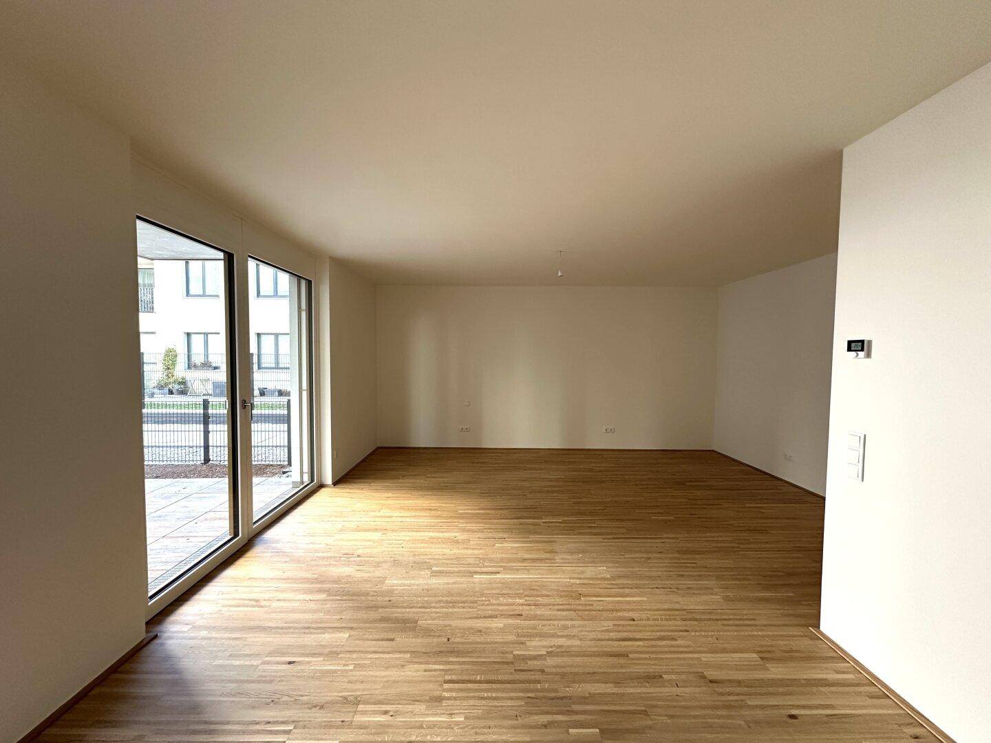 Heller, leerer Wohnraum mit Holzboden und großen Fenstern zum Außenbereich.