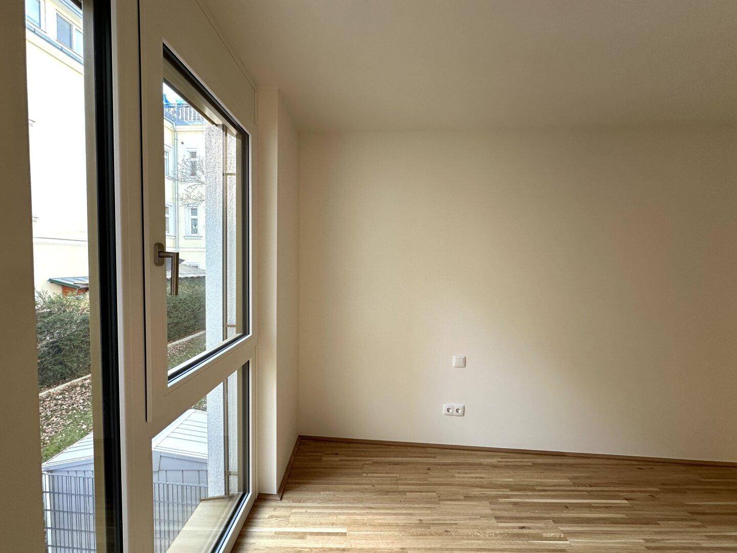Helles Zimmer mit Parkettboden und großem Fenster, das viel Tageslicht hereinlässt.