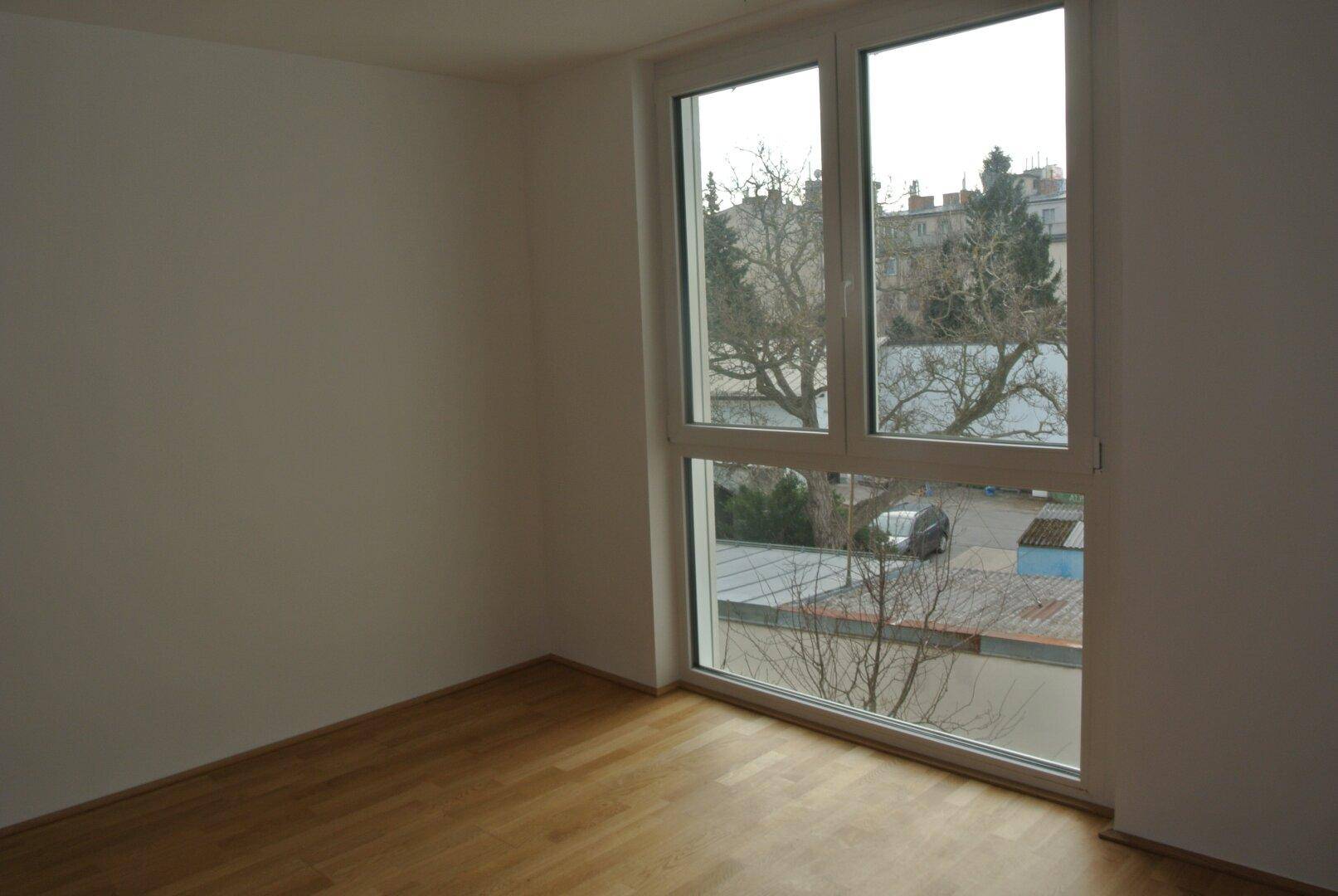 Heller, leerer Raum mit Holzparkettboden und großem Fenster mit Blick nach draußen.