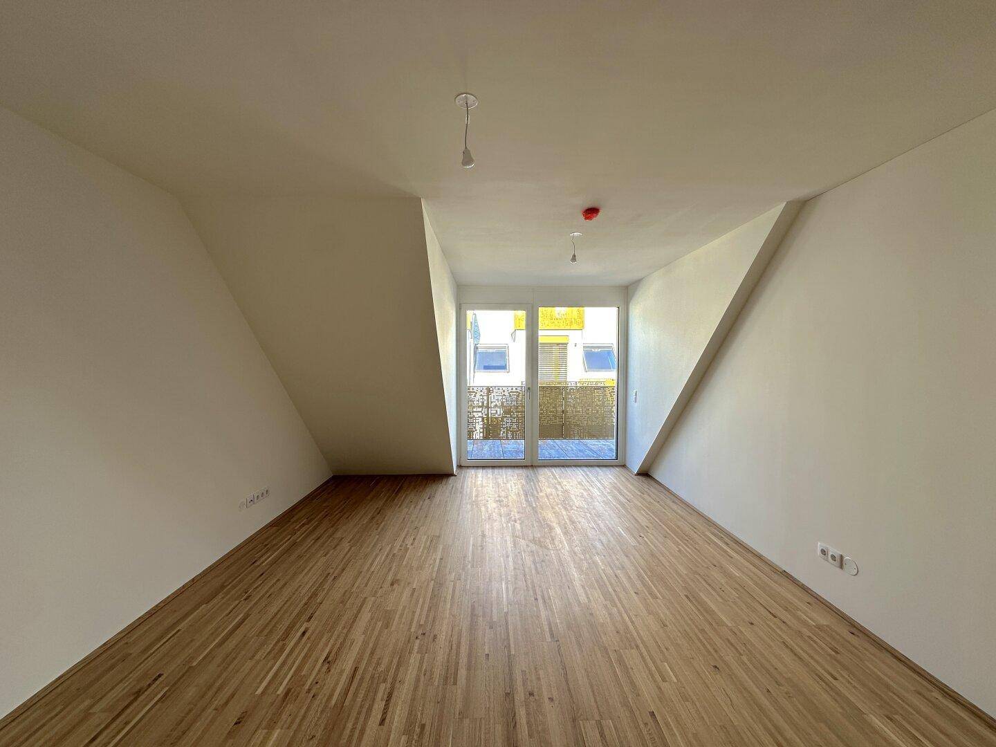 Geräumiger, heller Raum mit Holzboden, schrägen Wänden und Zugang zu einem Außenbereich.
