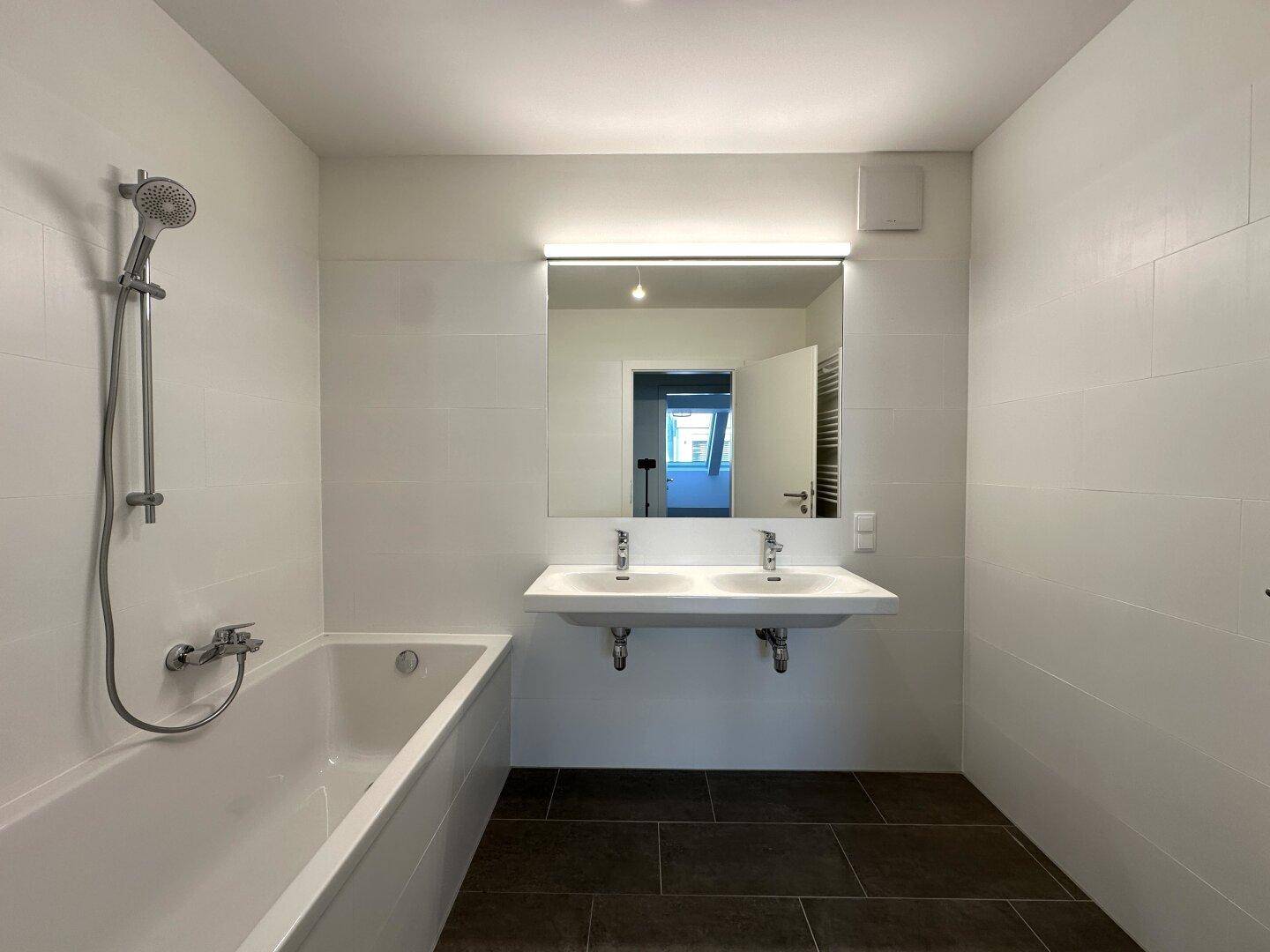 Modernes Badezimmer mit Badewanne, Doppelwaschbecken und großem Spiegel, hell gefliest.